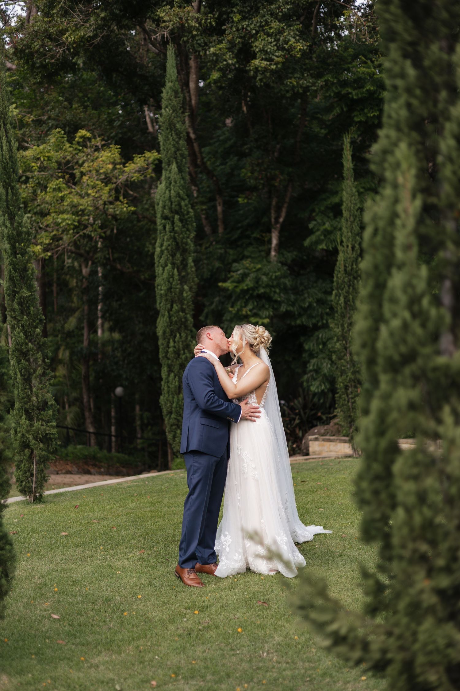 Bride and groom share a romantic kiss surrounded by tall cypress trees and lush greenery in a garden setting.