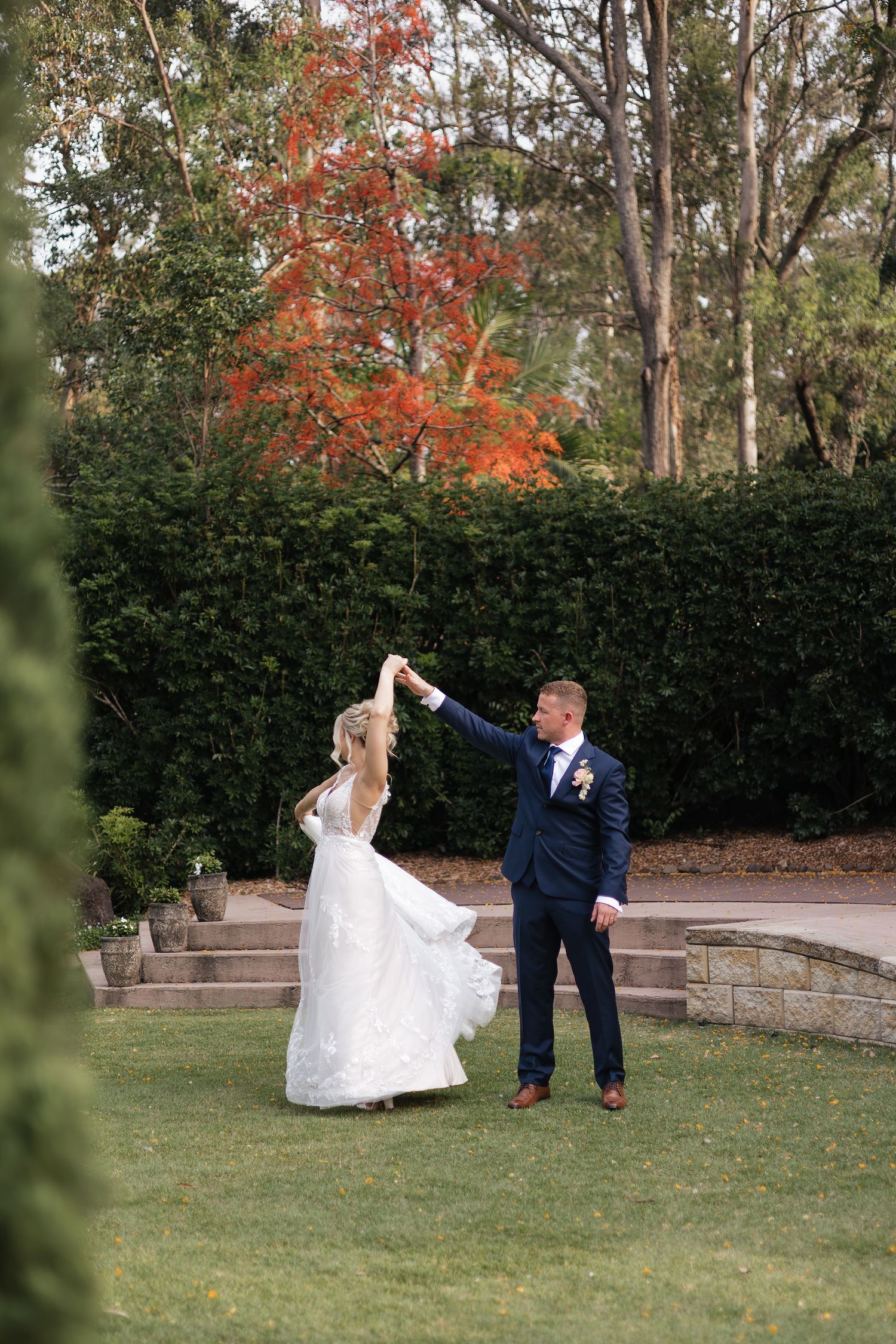 Groom twirling bride in white gown on garden lawn, with vibrant red autumn tree and green hedges backdrop.