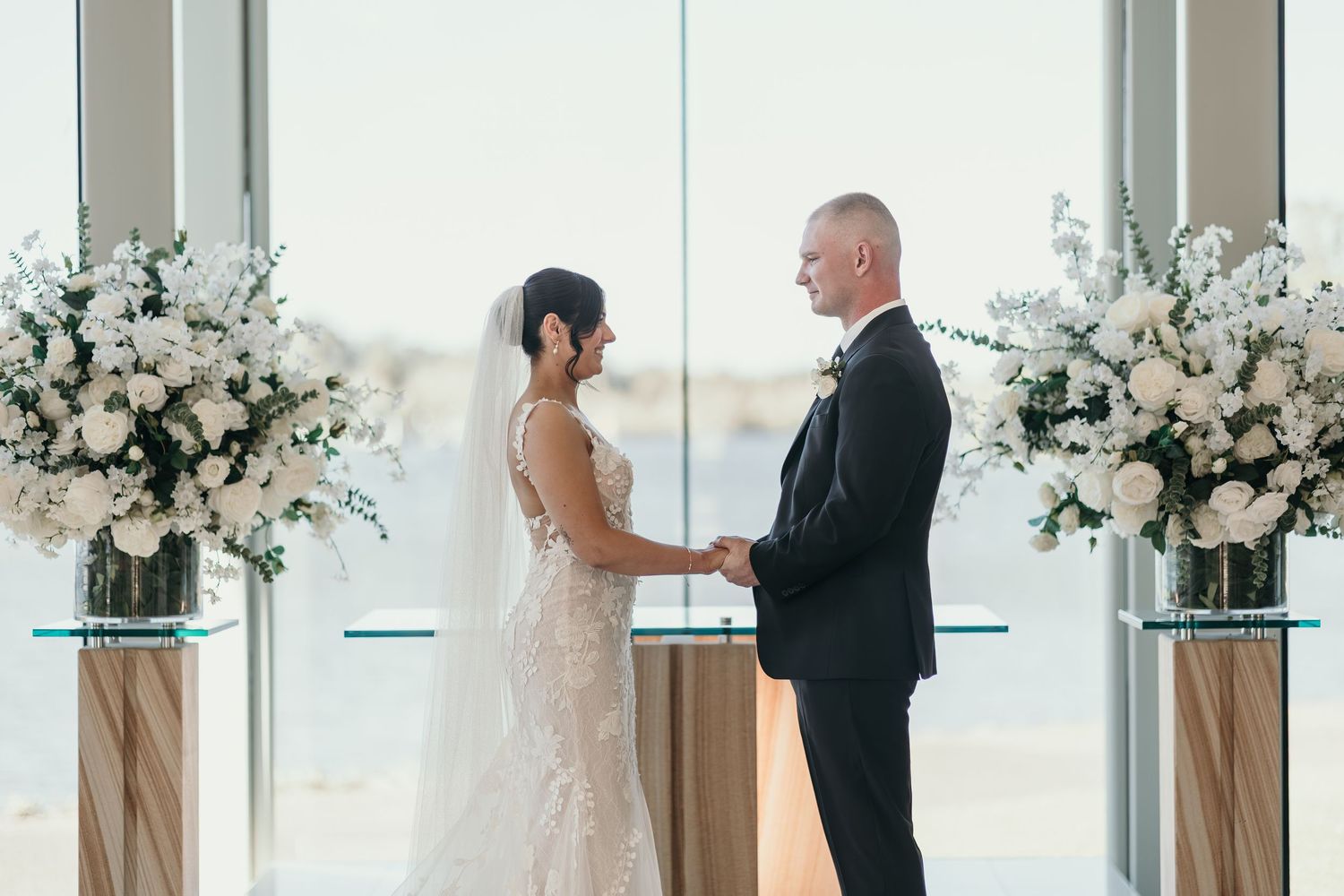 A couple exchanges vows during an elegant indoor wedding ceremony surrounded by white floral arrangements.