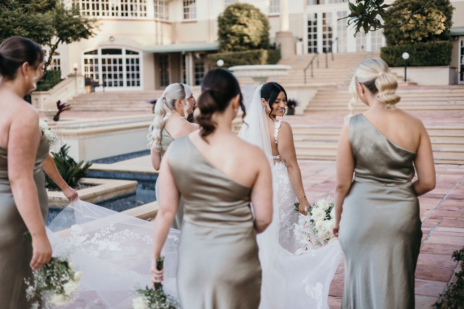 Bridesmaids in elegant champagne-colored satin dresses gather on stone steps holding delicate white bouquets.