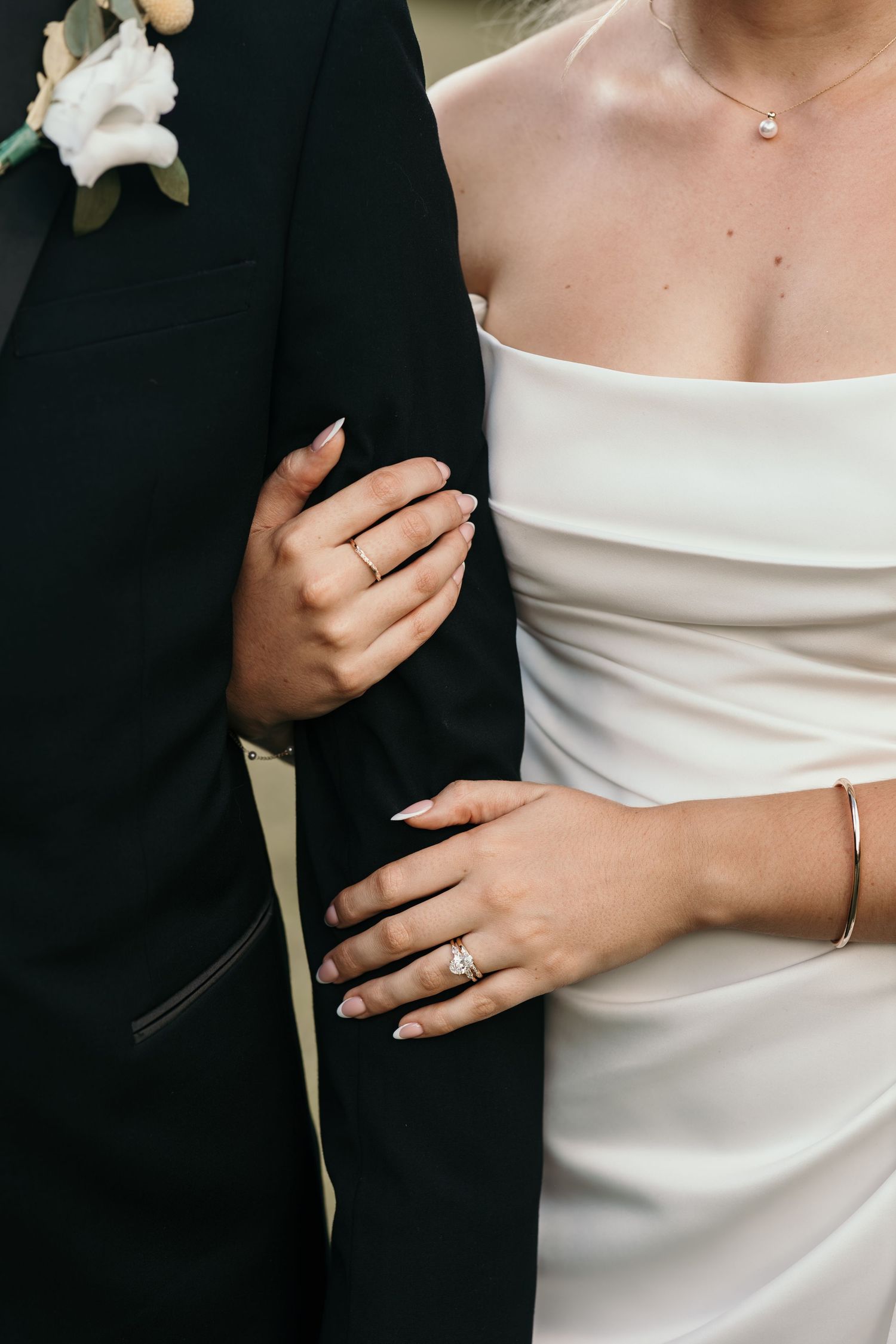 Bride and groom embrace, showcasing wedding rings, a diamond bracelet, and a white strapless gown with a delicate necklace.