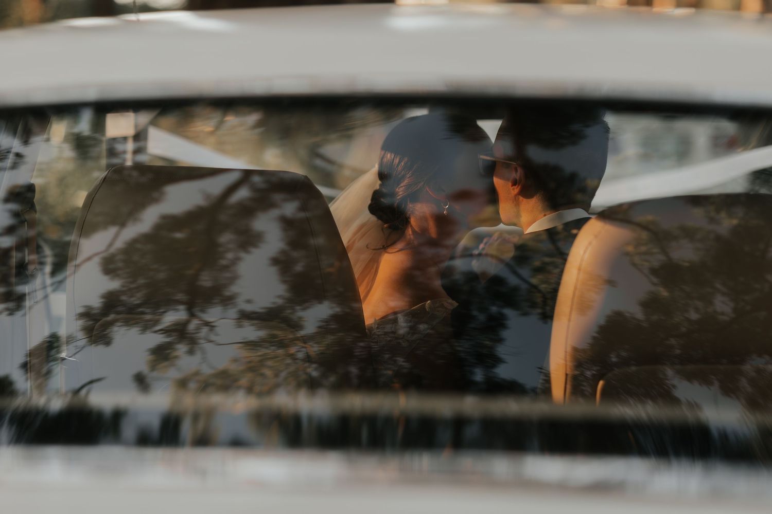 Couple sharing a romantic moment inside a car, captured through the windshield with tree reflections.