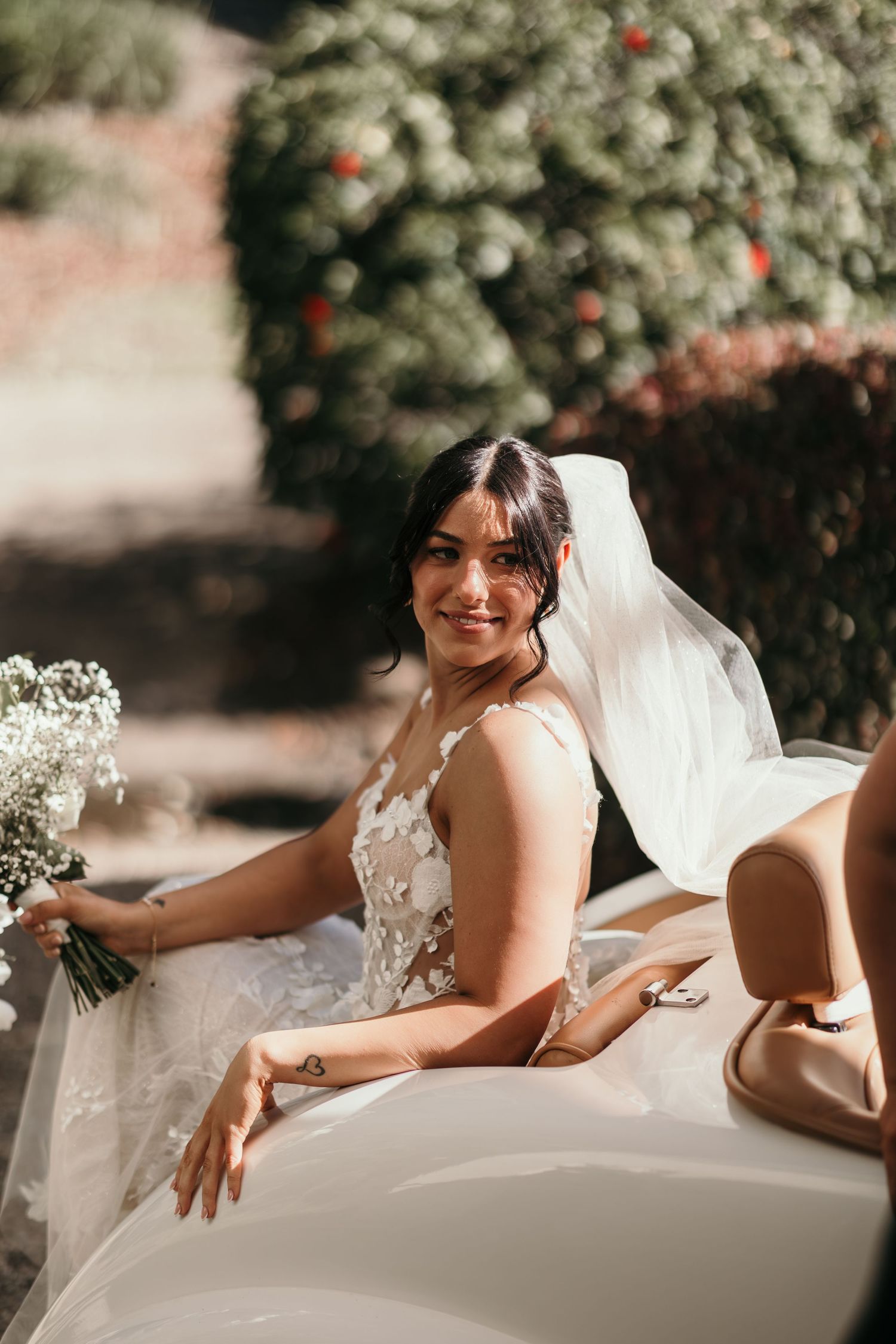 A bride in a white lace wedding dress poses elegantly on a chaise lounge outdoors with flowering trees in the background.
