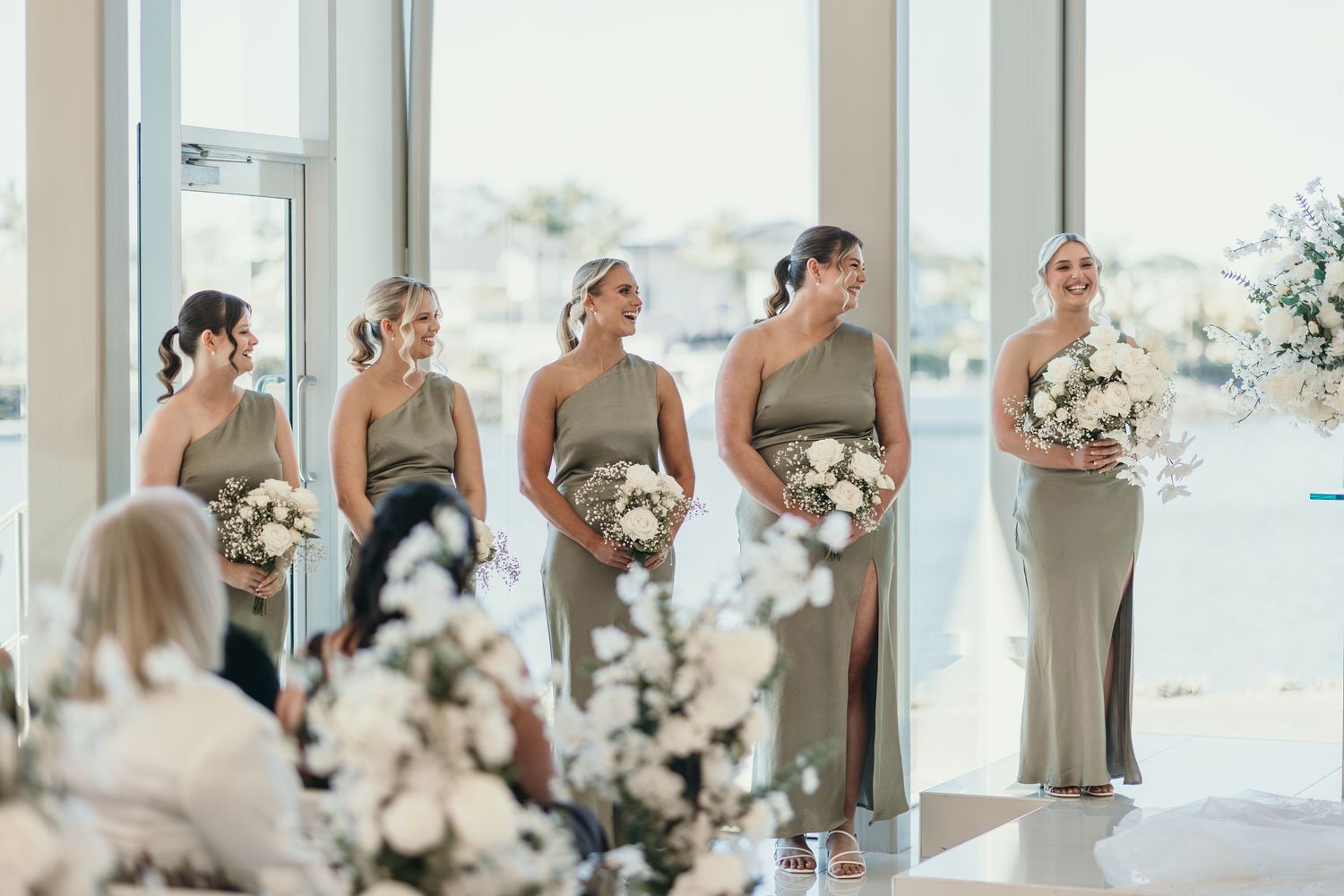A row of bridesmaids in sage green dresses stand in line looking out windows in an elegant venue.