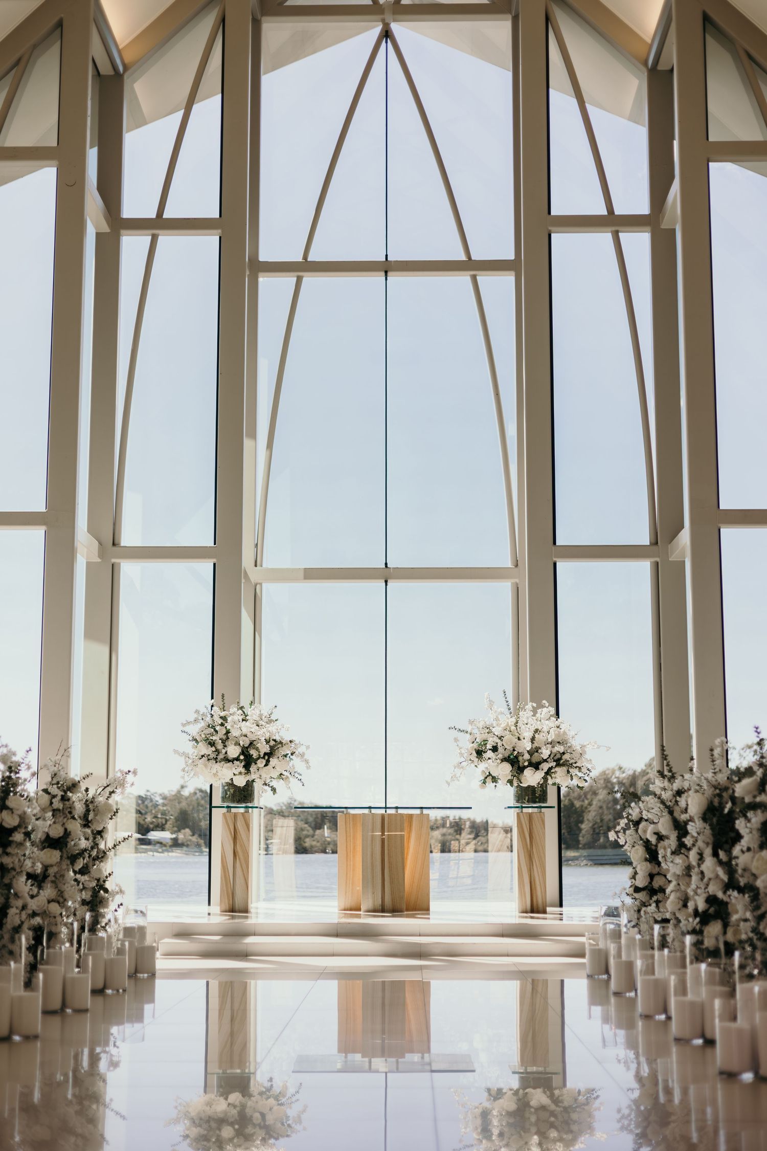 Modern white wedding chapel interior with floor-to-ceiling windows overlooking the ocean and white floral arrangements lining the aisle.