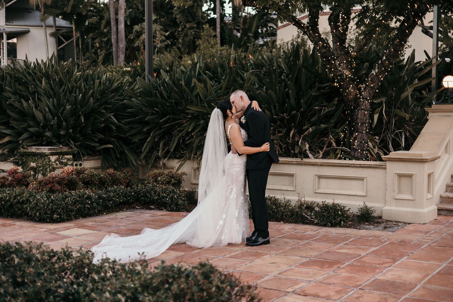 A romantic wedding couple embraces in a lush garden setting with terracotta tiles and greenery.