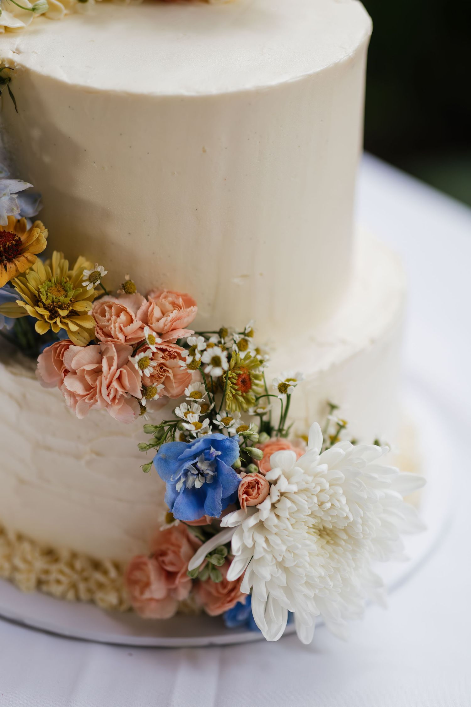 Two-tier white buttercream wedding cake decorated with fresh pink, blue, and white flowers including chrysanthemums.
