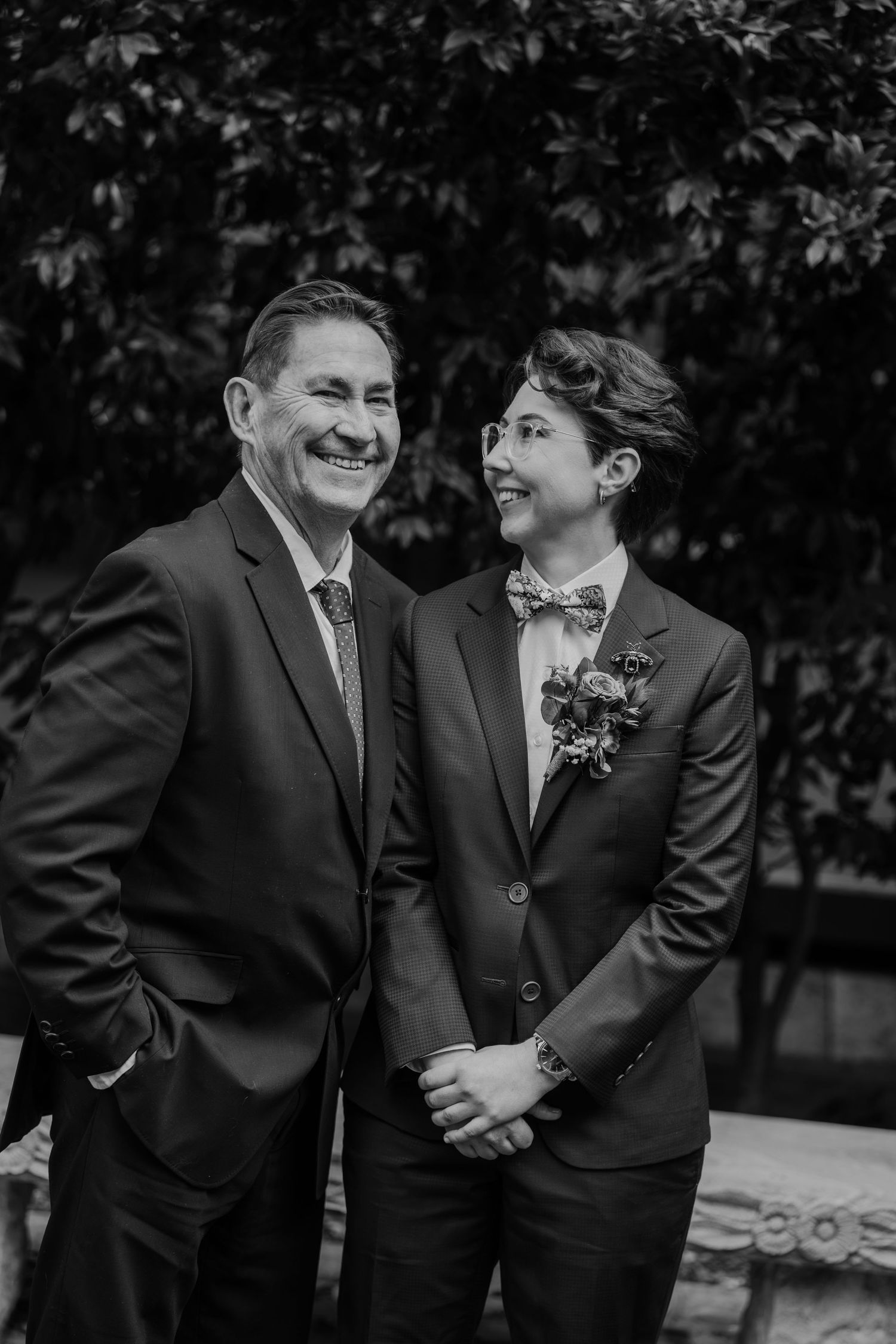 Black and white photo of two people in suits smiling at a wedding, one wearing a bow tie and floral boutonniere.