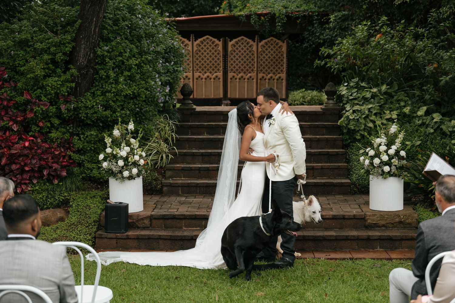 Bride and groom share first kiss at outdoor wedding ceremony with floral arrangements, stone steps, and dogs nearby.