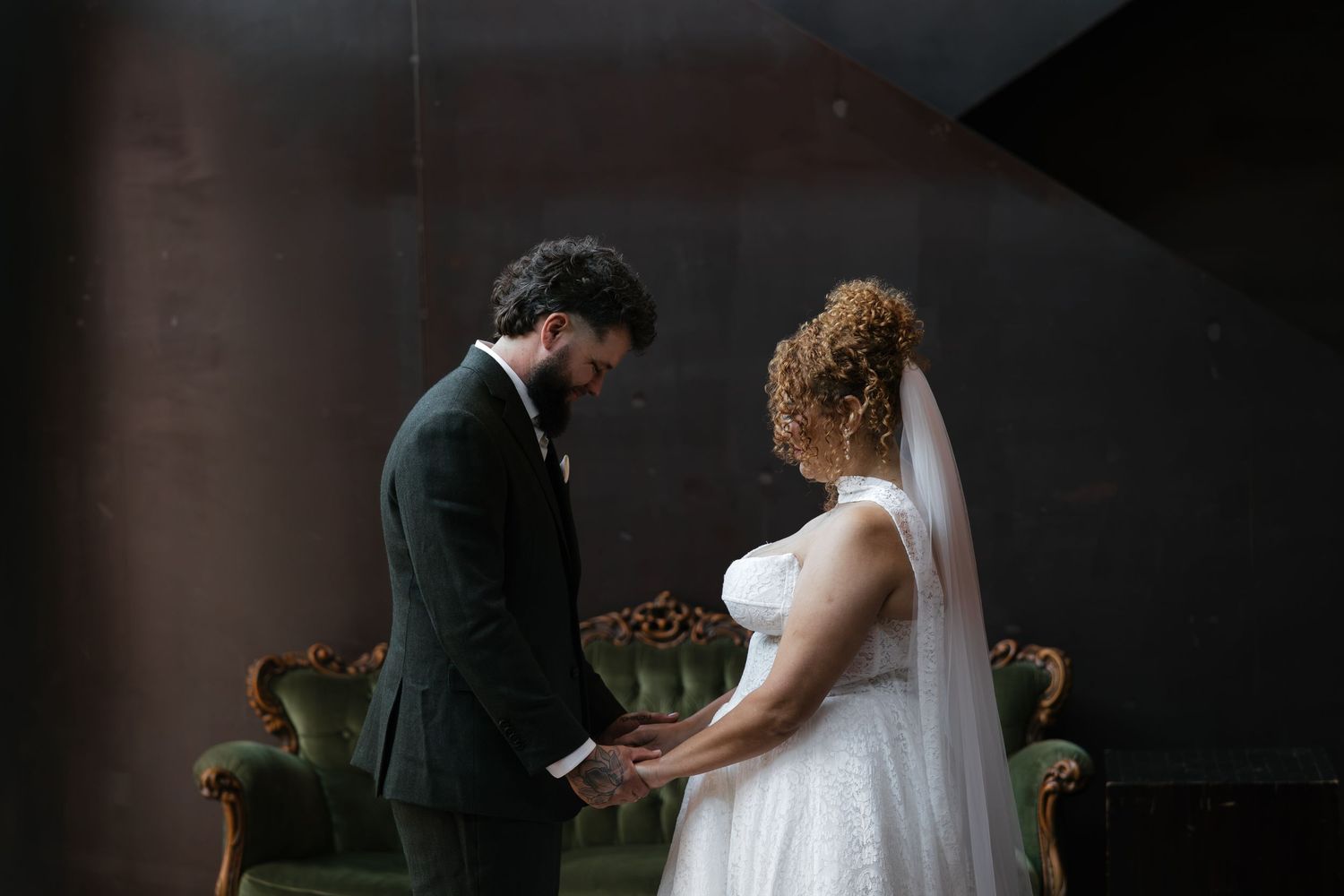 Bride and groom holding hands during intimate wedding ceremony in elegant dark venue with green velvet chairs.