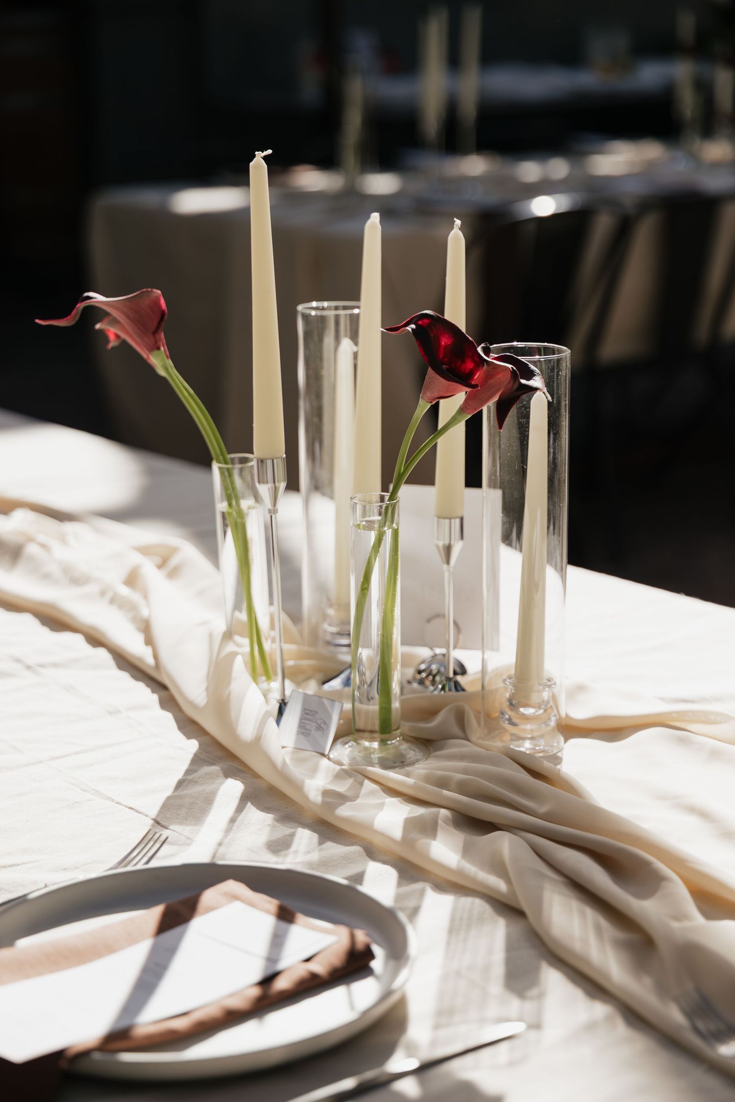 Elegant wedding table setting with red calla lilies, tall white candles in glass holders, and neutral linen runner.