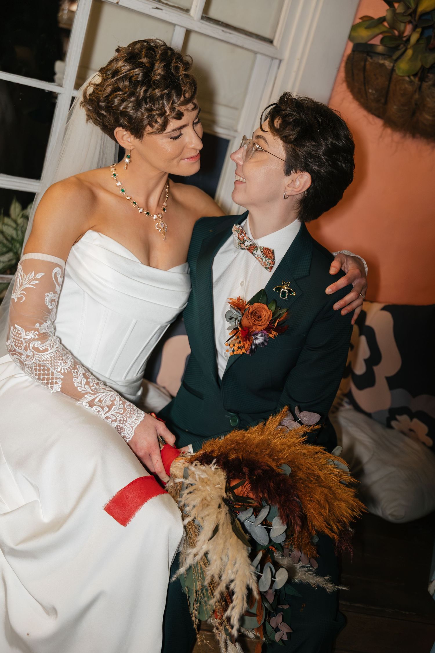 Two brides in wedding attire share a loving gaze, one in white gown with lace sleeves, other in green suit with floral boutonniere.