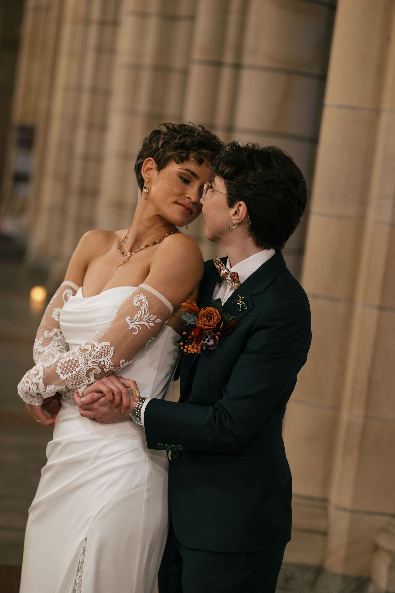 Two brides in wedding attire share a romantic moment, one in a white lace gown, the other in a dark suit with floral boutonniere.