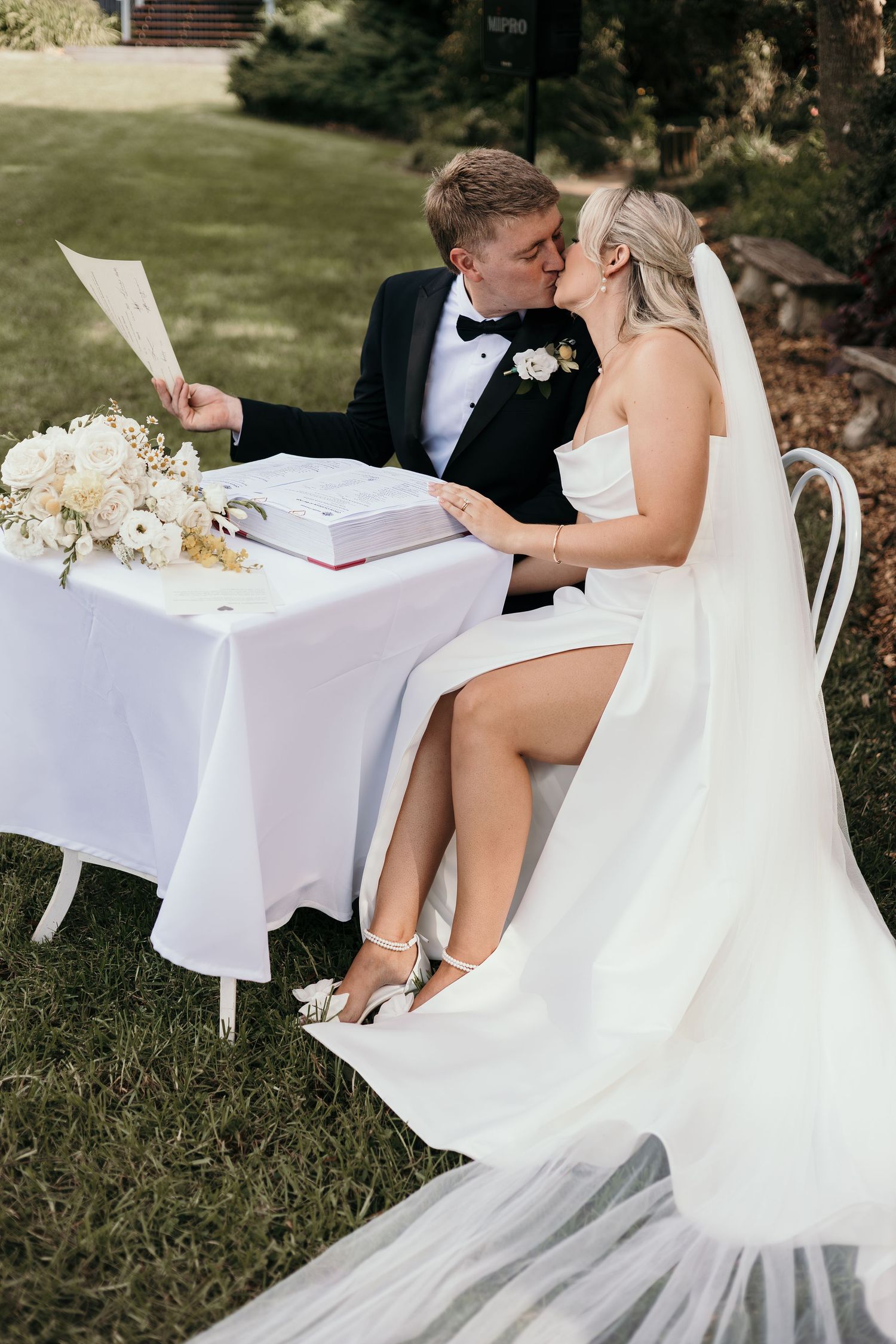 Bride and groom share a kiss while seated at a signing table outdoors, surrounded by white floral arrangements.