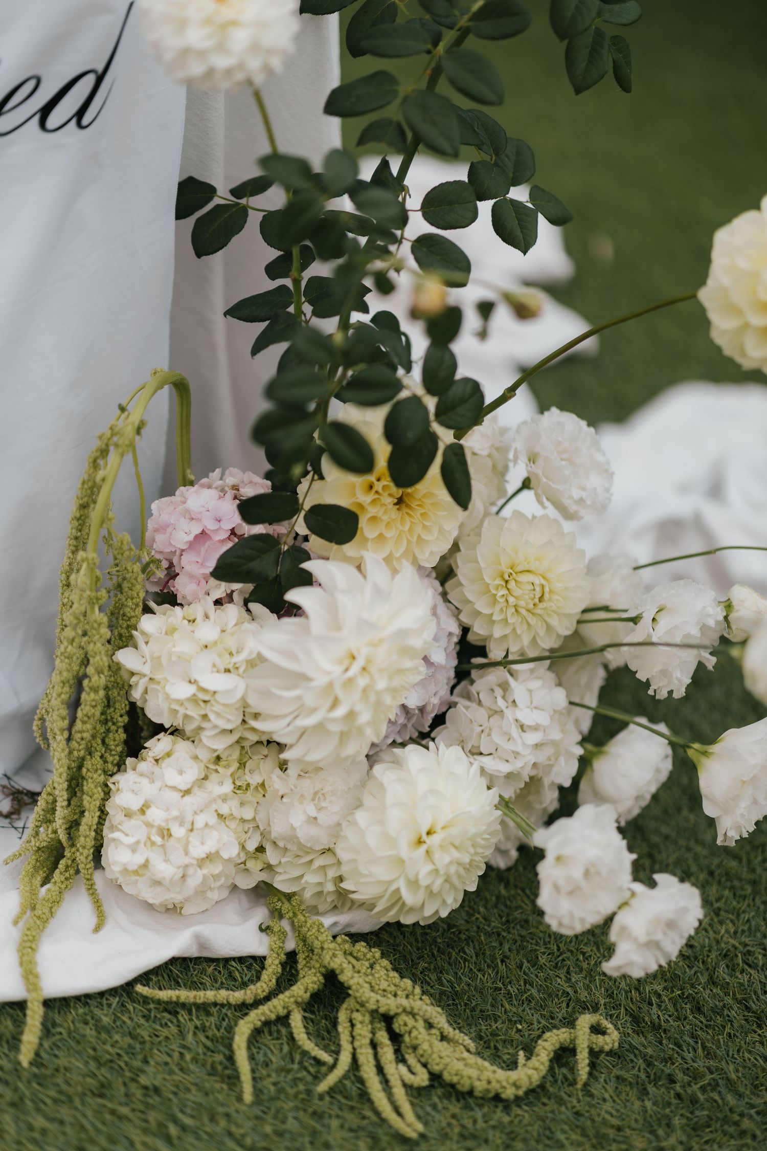 White dahlias and hydrangeas with green eucalyptus branches arranged on grass at an outdoor wedding ceremony.