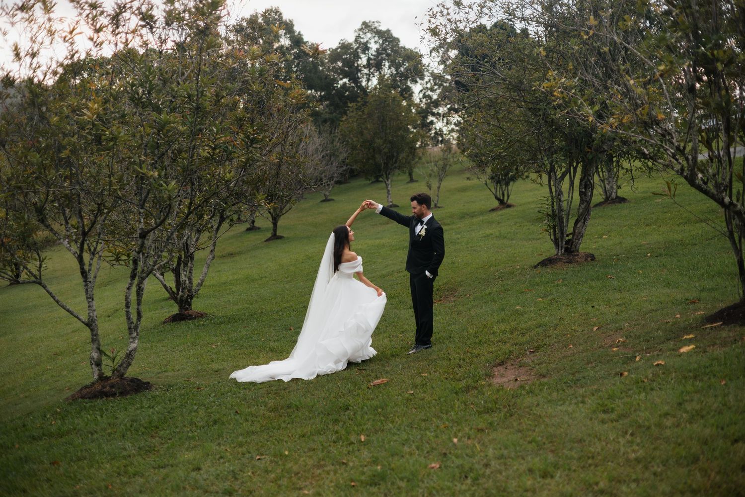 Bride and groom dancing outdoors in a lush green orchard, bride in flowing white gown, groom in black suit.