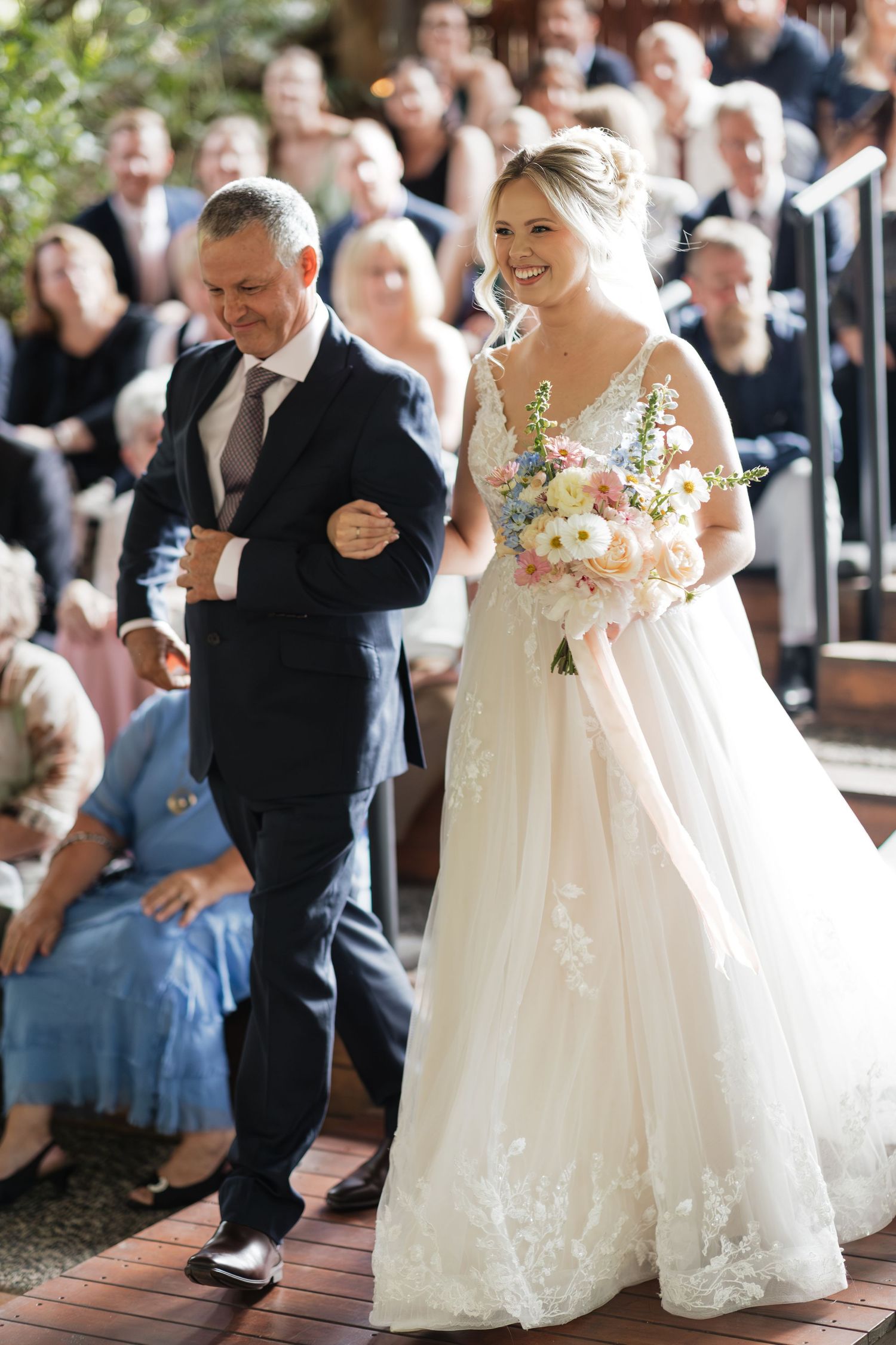 Bride in floral lace gown holding pastel bouquet walks down aisle with escort at outdoor wedding ceremony.