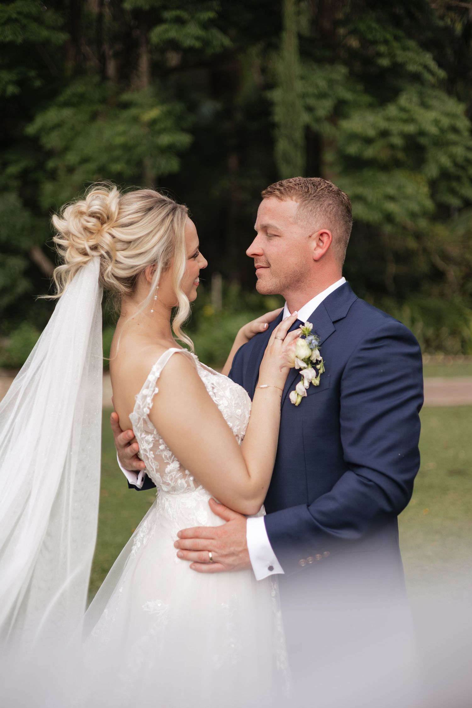 Bride and groom share an intimate moment outdoors, with bride adjusting grooms navy suit tie while smiling.