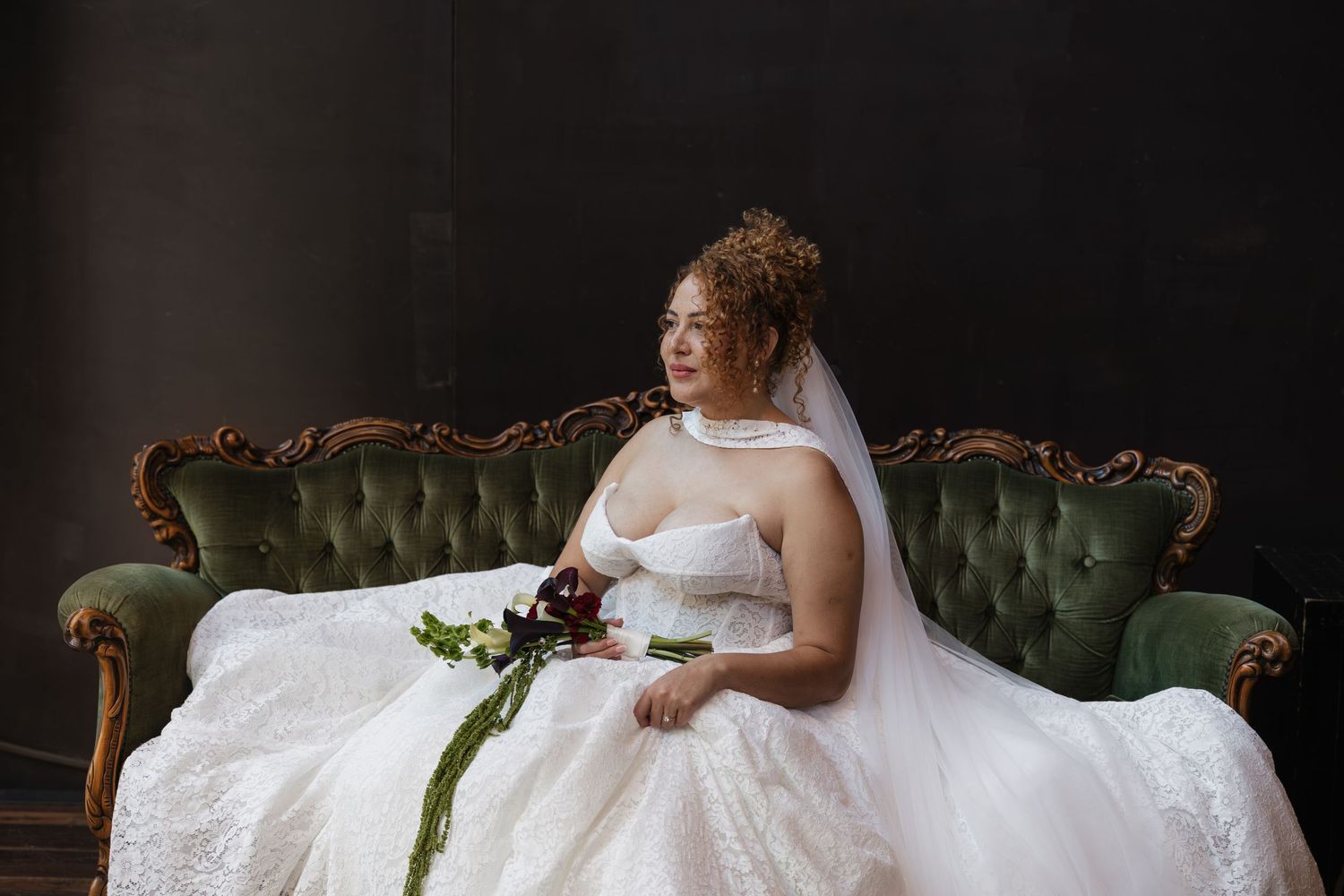 Bride in white lace gown and veil sits on green velvet sofa holding bouquet, gazing away in elegant dark studio.
