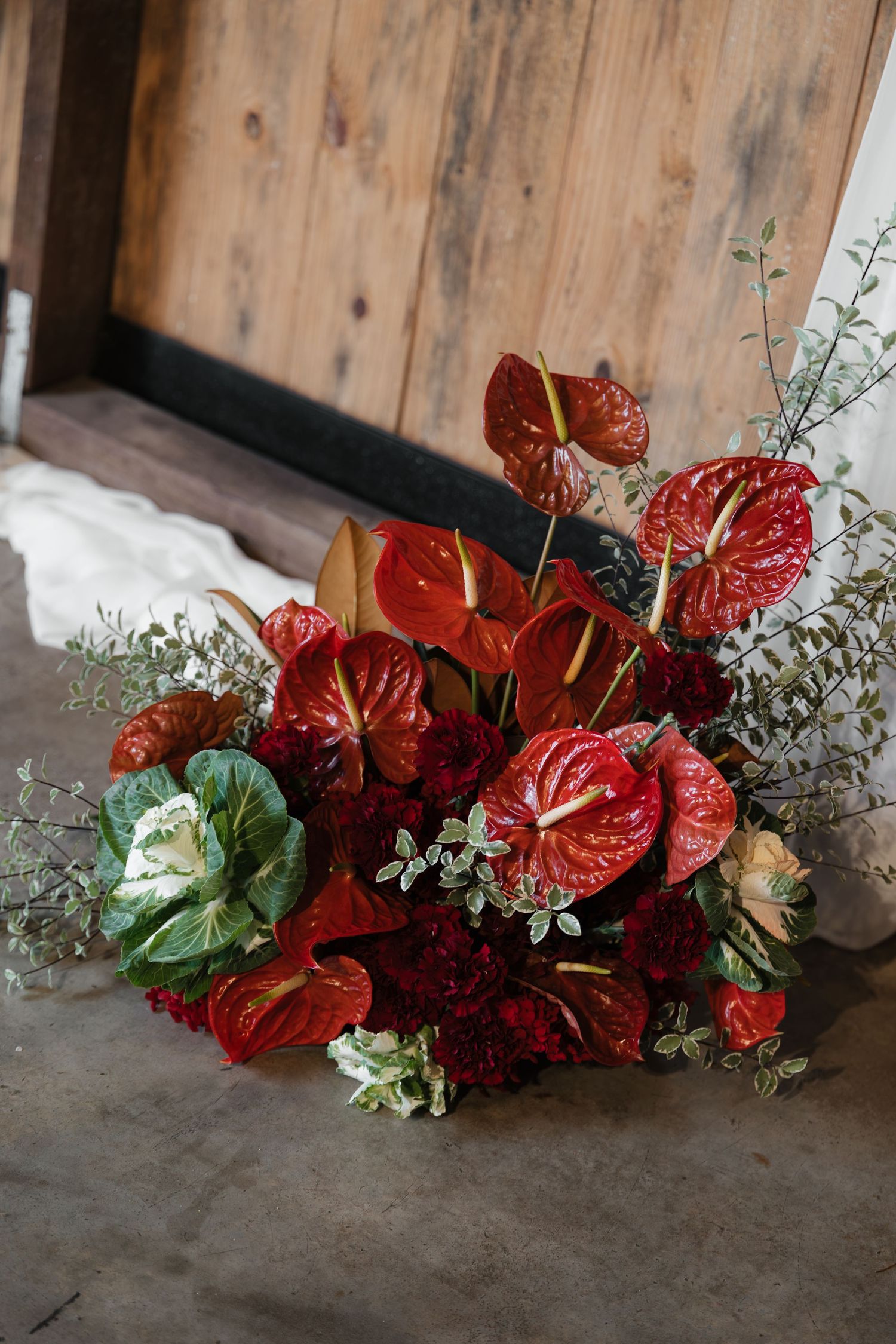Rich red anthurium bouquet with dark blooms and greenery displayed on concrete floor near wooden door.