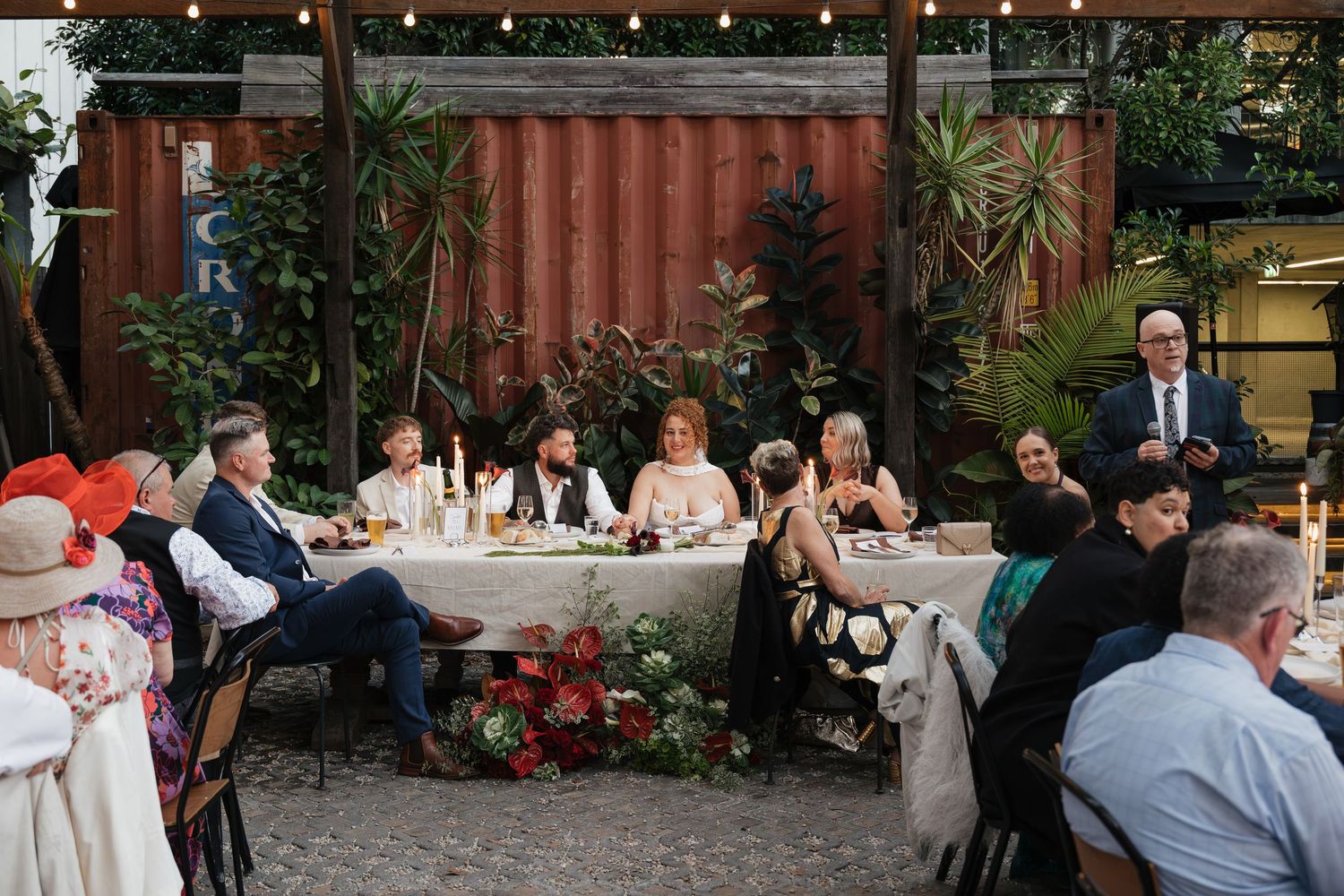Guests seated at an elegant outdoor wedding reception table with floral centerpieces, candles, and a man giving a speech.