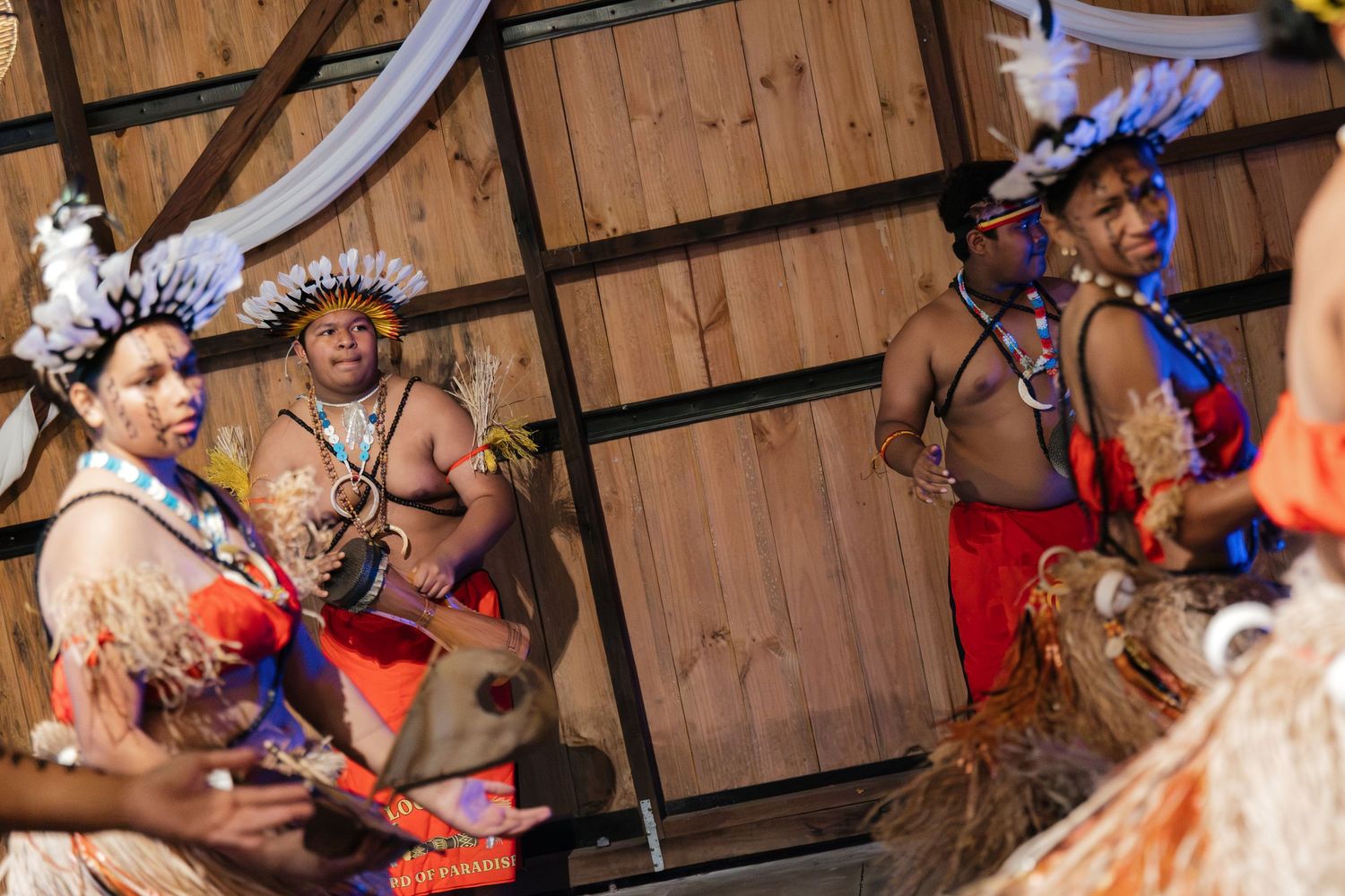 Traditional Pacific Islander dancers in colorful ceremonial costumes performing a cultural dance ceremony indoors.