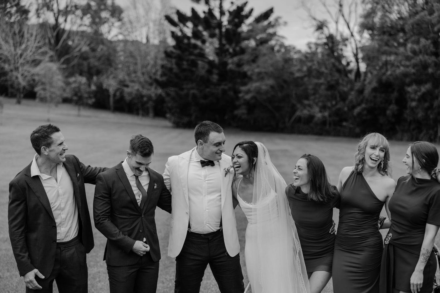Black and white wedding party photo with bride and groom surrounded by smiling guests on lush green grounds.