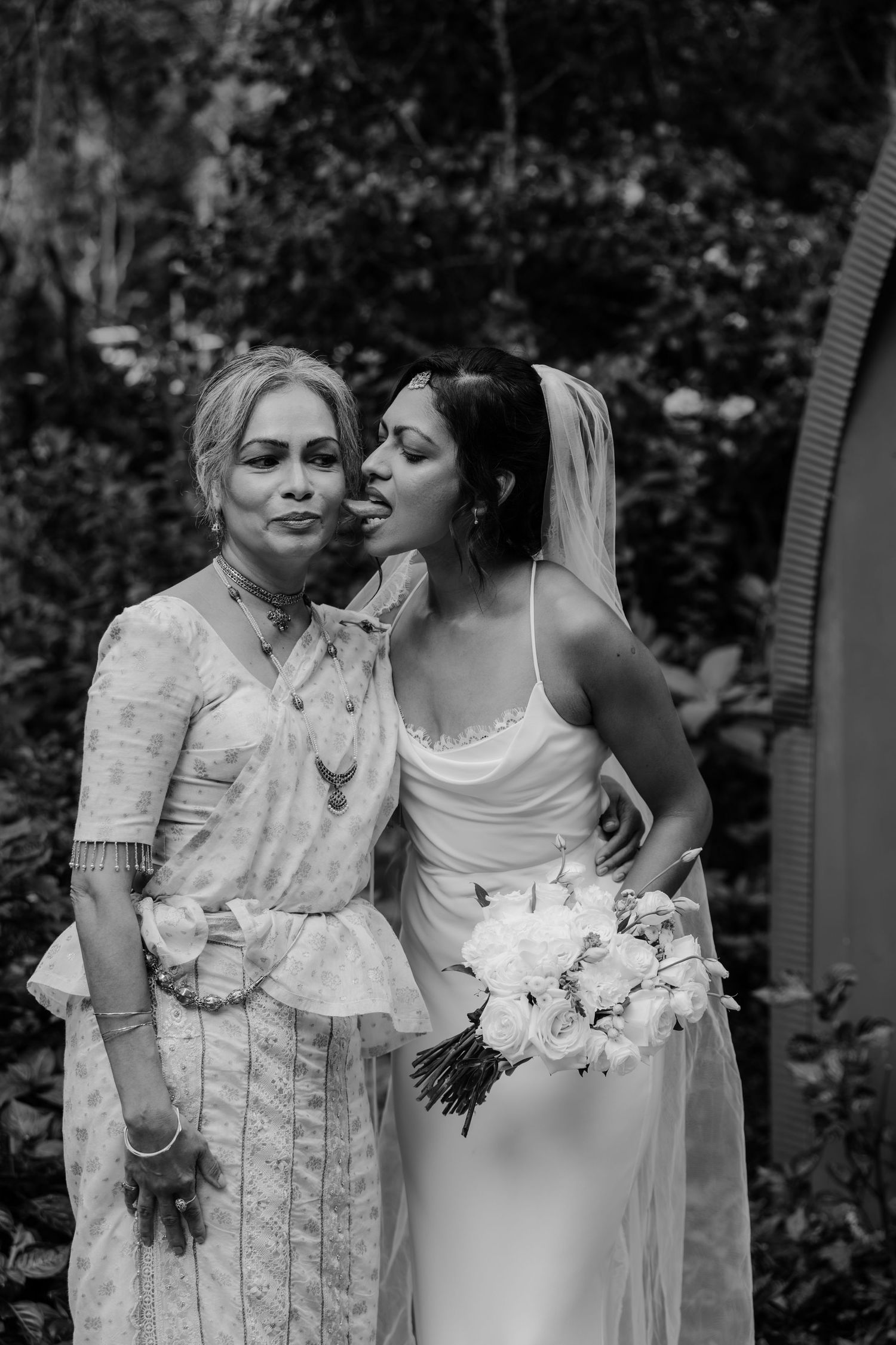 Black and white photo of a bride in a white gown and veil holding flowers, posing with an older woman in traditional attire.