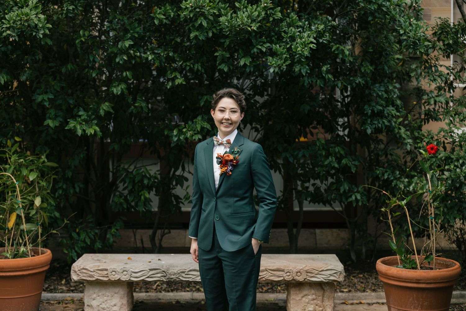 Person in dark green suit with floral boutonniere standing in garden courtyard with terracotta pots and lush greenery.