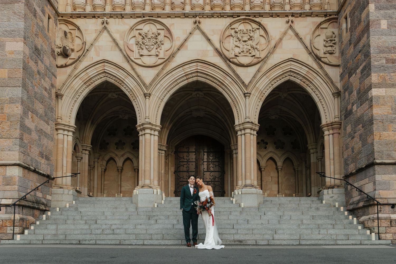 Bride and groom standing on stone steps before ornate Gothic cathedral arched entrance on wedding day.