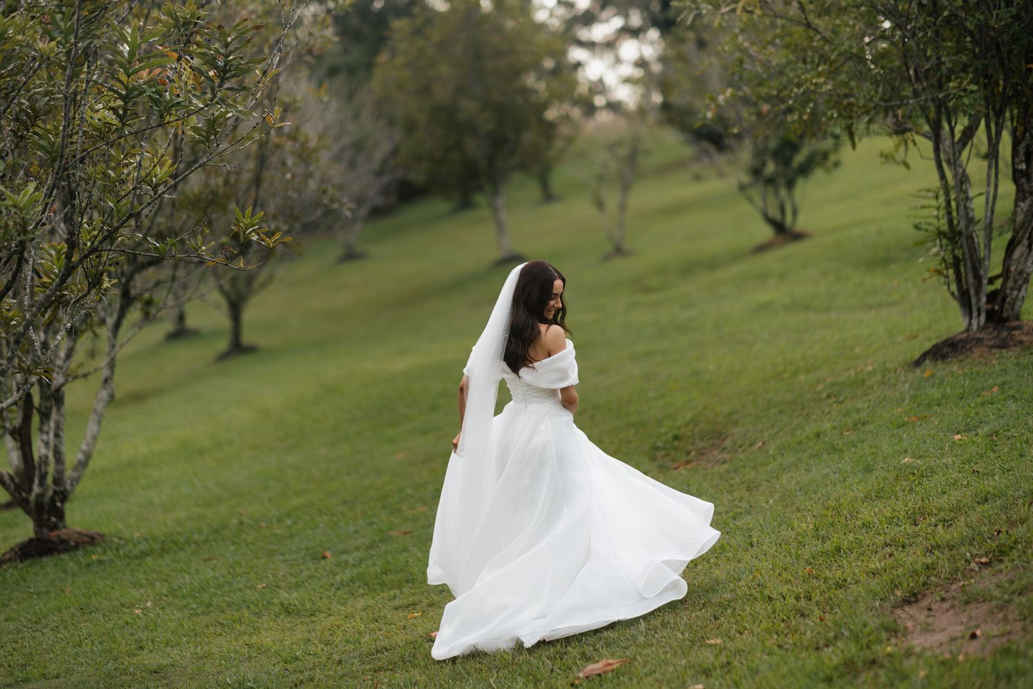Bride in white off-shoulder gown and veil walks through lush green orchard on her wedding day.
