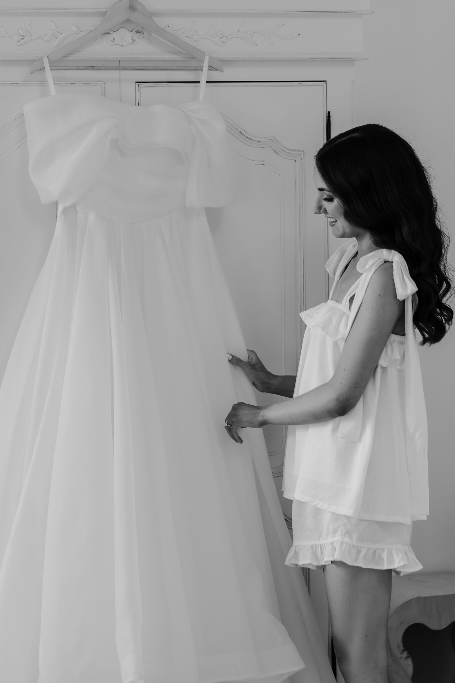 Black and white photo of a smiling bride in pajamas admiring her elegant wedding dress hanging on a door.