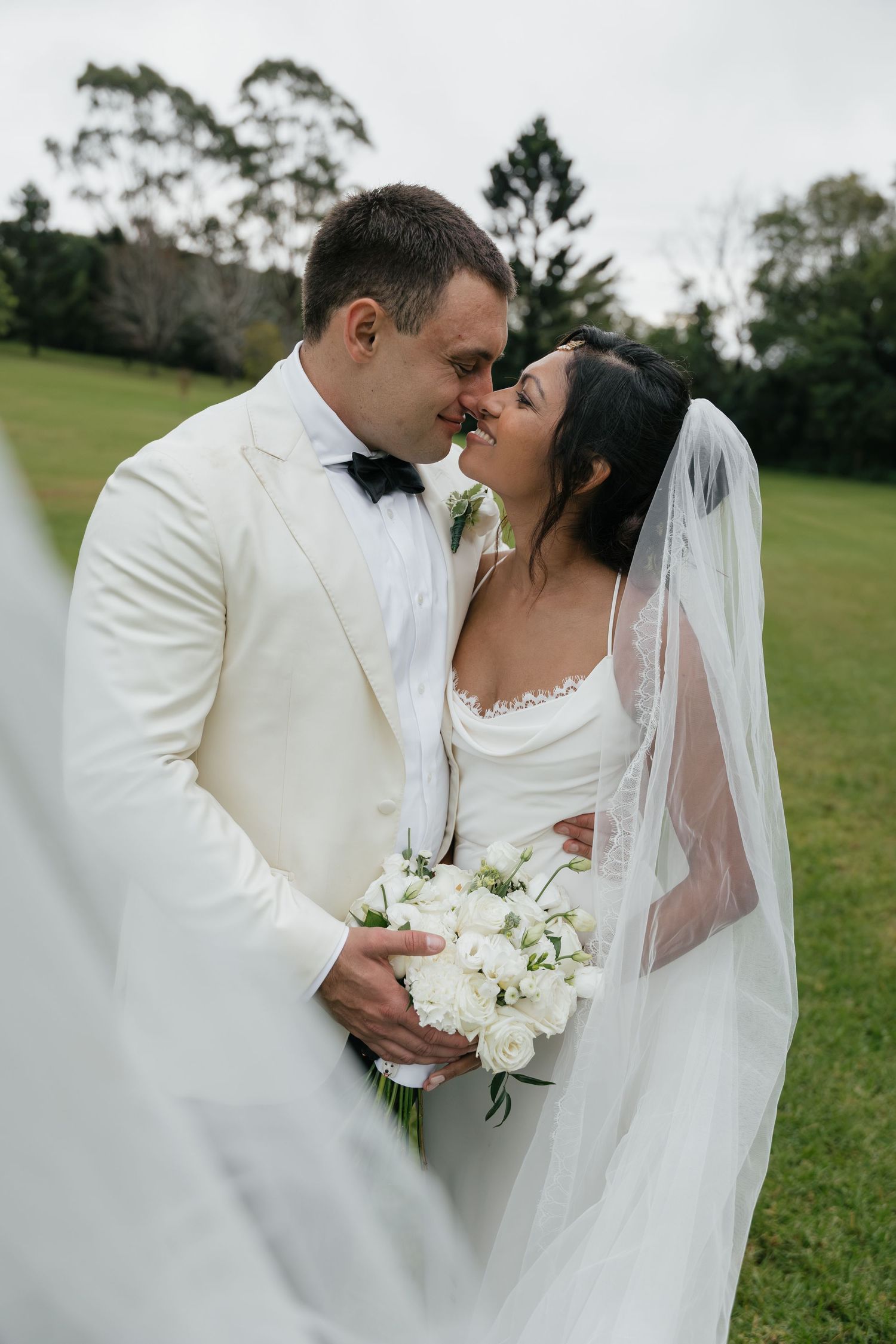 Bride and groom in white wedding attire share a romantic moment outdoors, with bride holding white rose bouquet and veil flowing.
