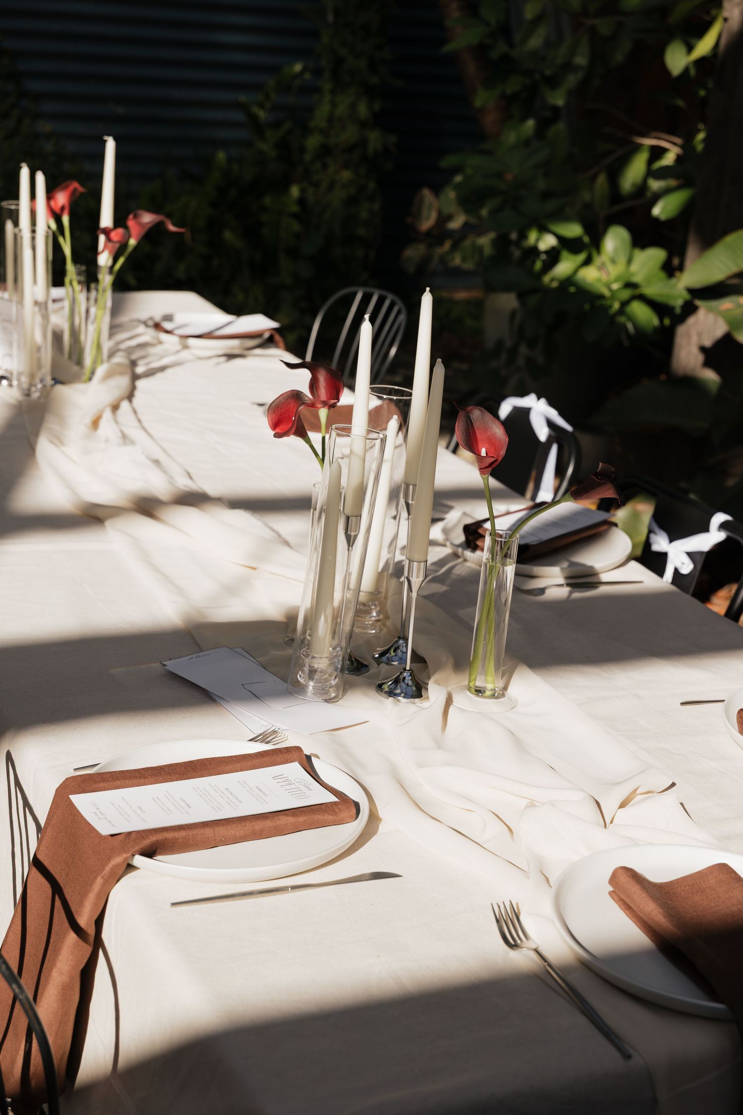 Elegant outdoor dining table set with white linen, taper candles, red flowers, and place settings in natural sunlight.