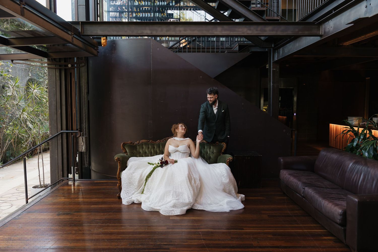 Bride in white gown seated on green velvet sofa with groom standing beside her in a modern industrial wedding venue.