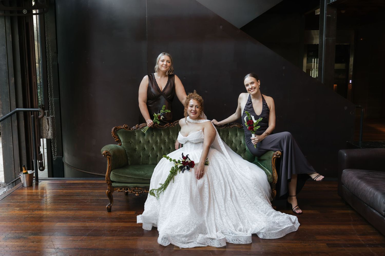 Bride in white gown sits on green velvet sofa flanked by two bridesmaids in dark dresses holding bouquets.