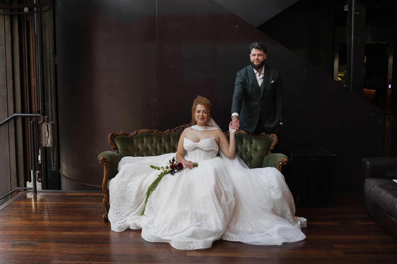 Bride in white ball gown sits on green velvet sofa holding groom's hand at elegant wedding venue.
