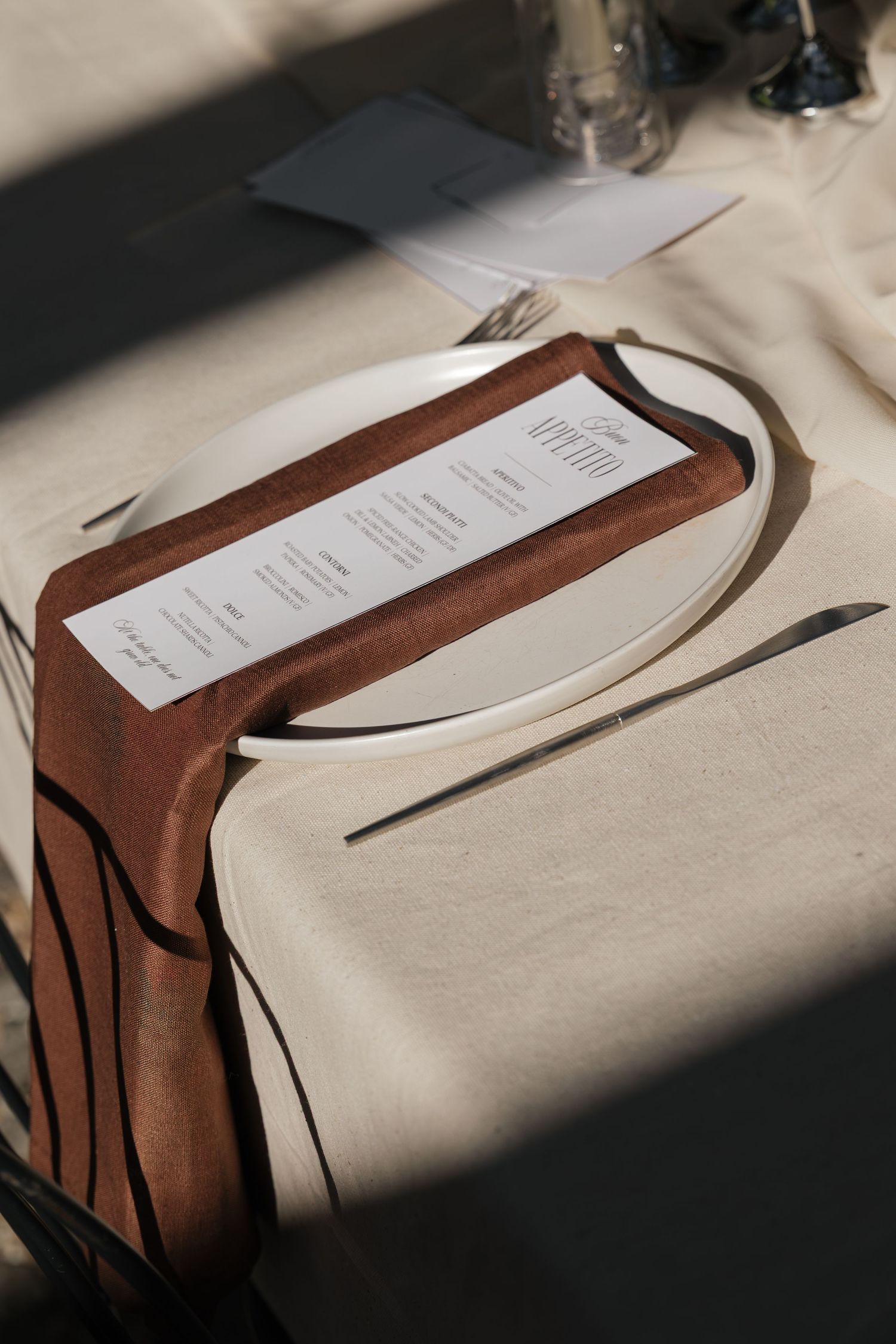 Elegant place setting with a white plate, brown linen napkin, and printed menu card on a neutral tablecloth.