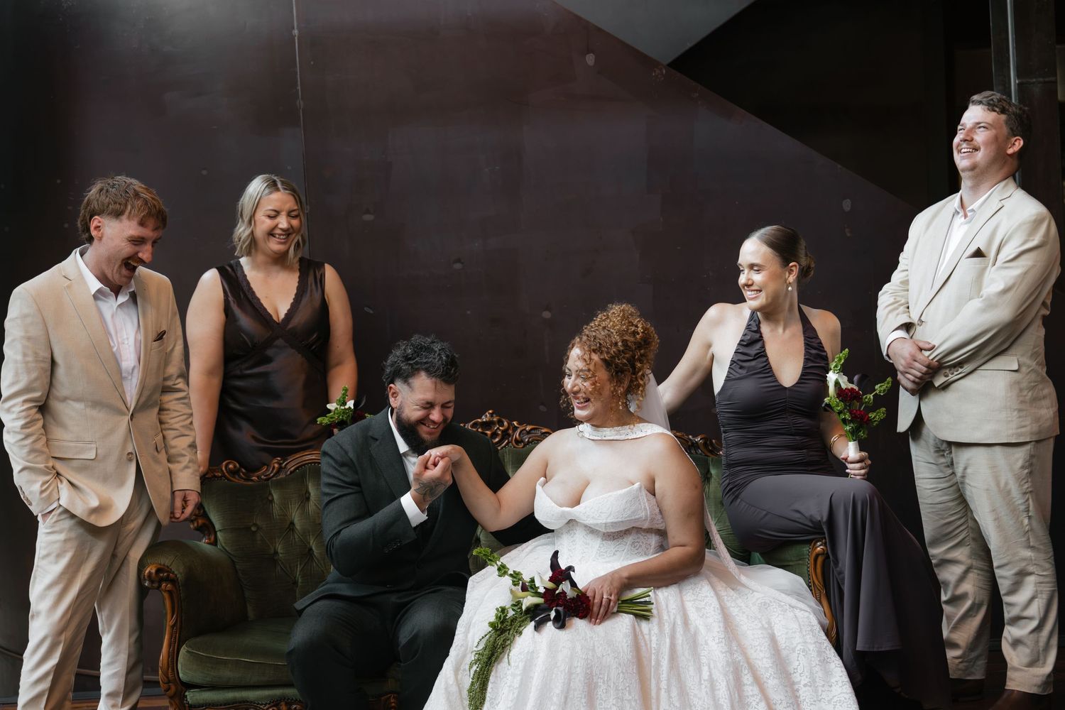 Wedding party laughing together, bride and groom seated while friends in black and neutral attire celebrate around them.
