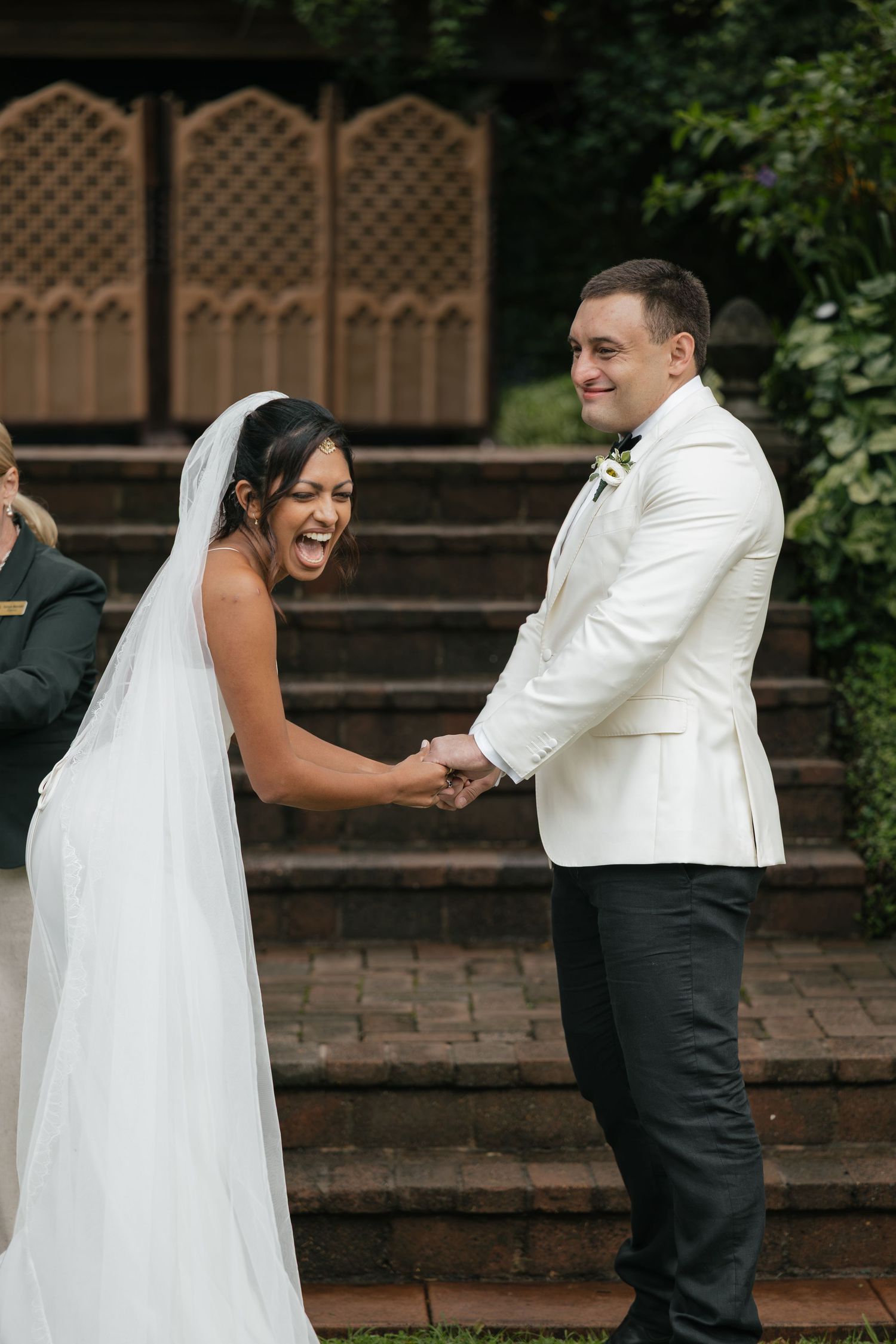 Bride laughing joyfully while holding hands with groom in white tuxedo jacket during outdoor wedding ceremony.