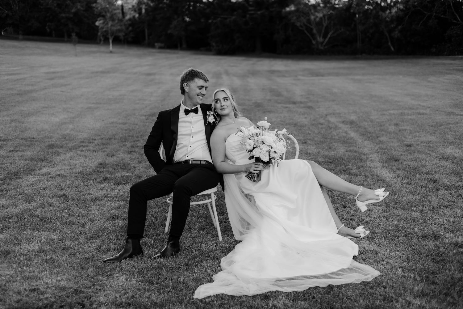 Black and white photo of a bride and groom sitting on grass, bride holding bouquet, groom in tuxedo.