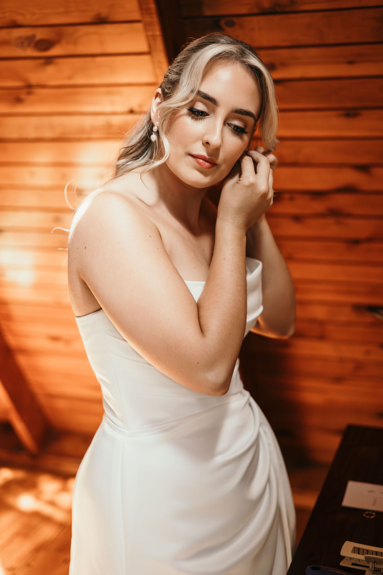 Blonde bride in strapless white gown adjusting earring in warm wooden cabin, elegant bridal portrait.