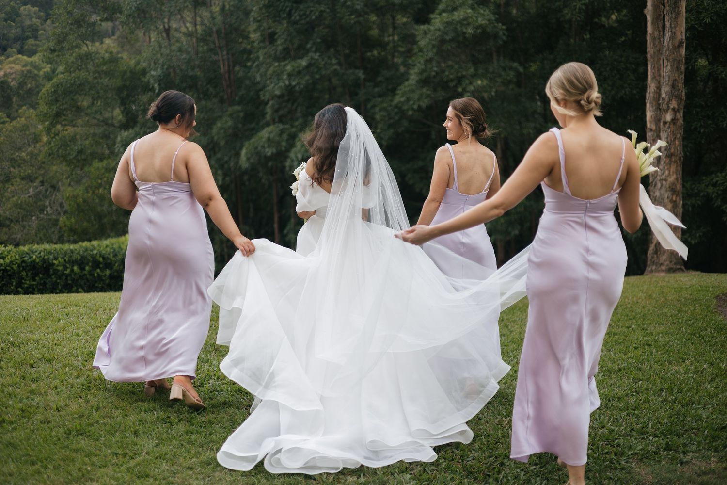 Bride in white gown and veil walks with three bridesmaids in lavender satin dresses on lush green lawn.