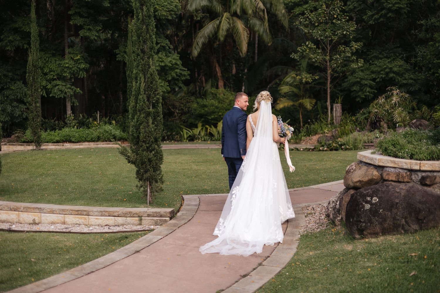 Bride in white gown and groom in navy suit walk along garden path surrounded by lush greenery on their wedding day.