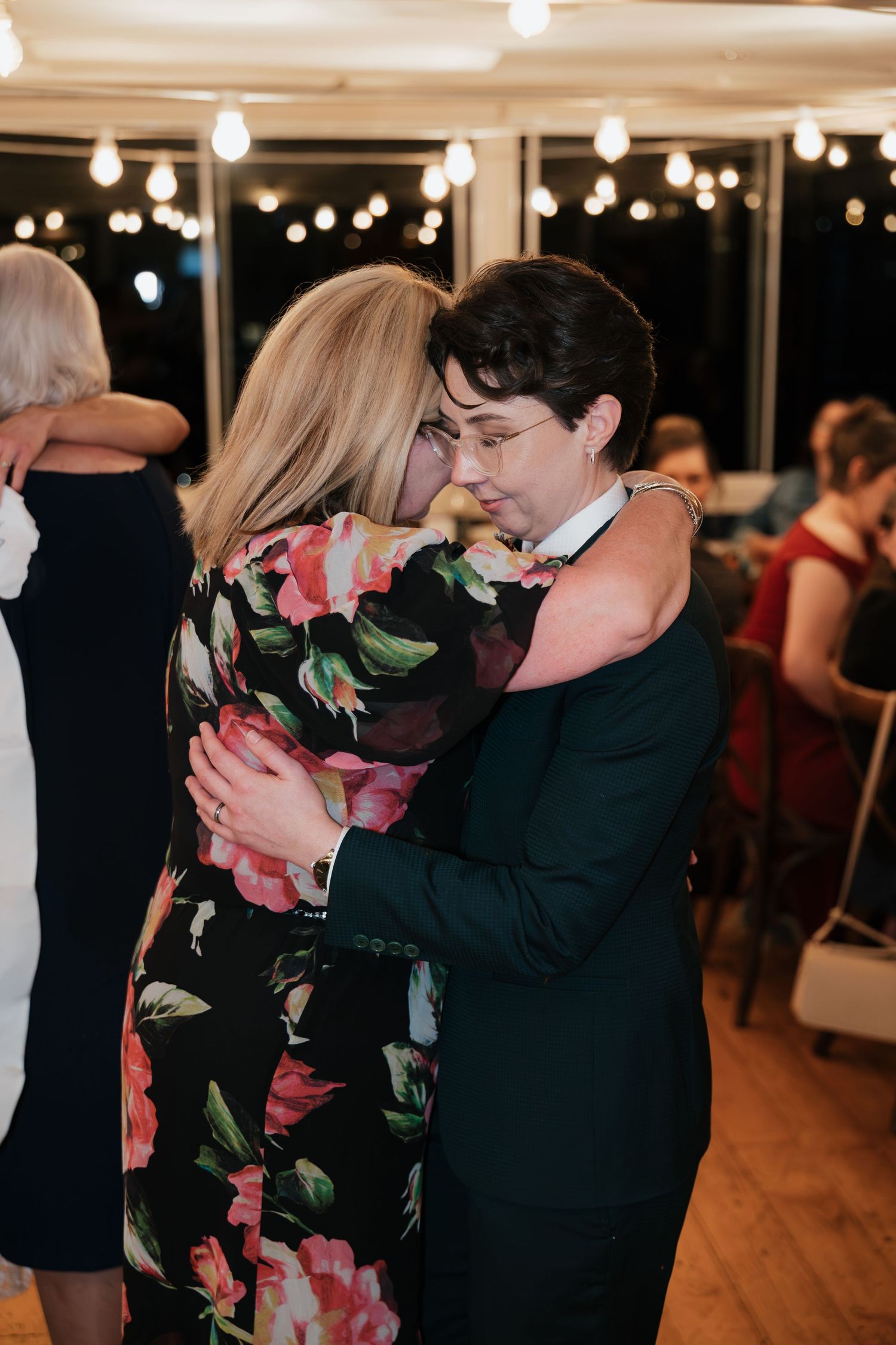 Two women share an emotional dance floor embrace at a wedding reception under warm string lights.