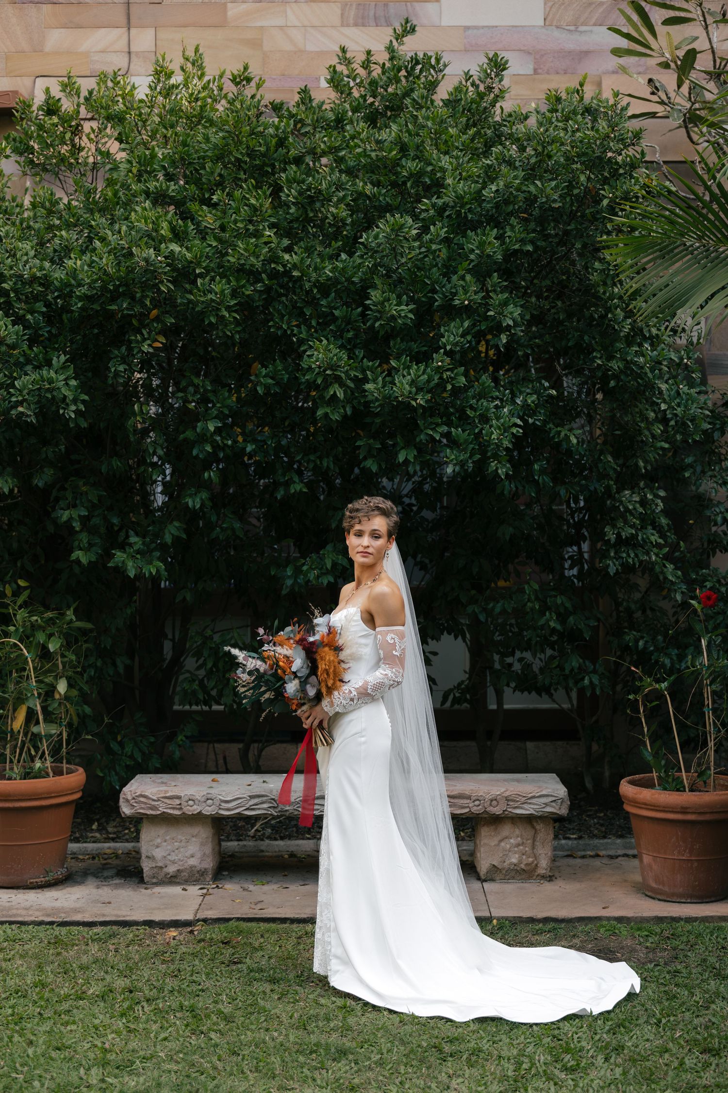 Bride in white lace gown with veil holds bouquet, standing before lush green trees and stone bench outdoors.