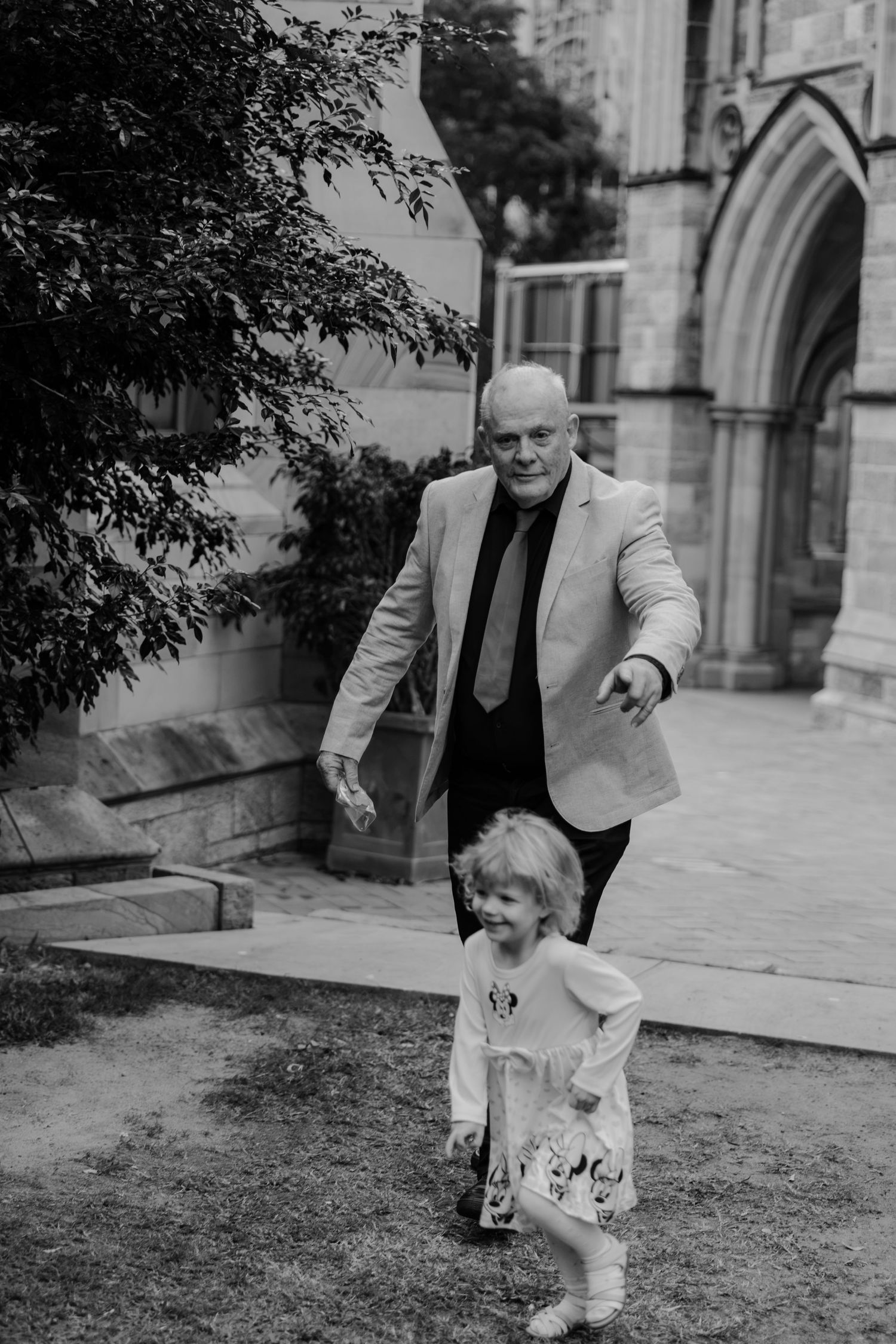 Black and white photo of a bald man in a suit jacket chasing a toddler girl outside a stone church.