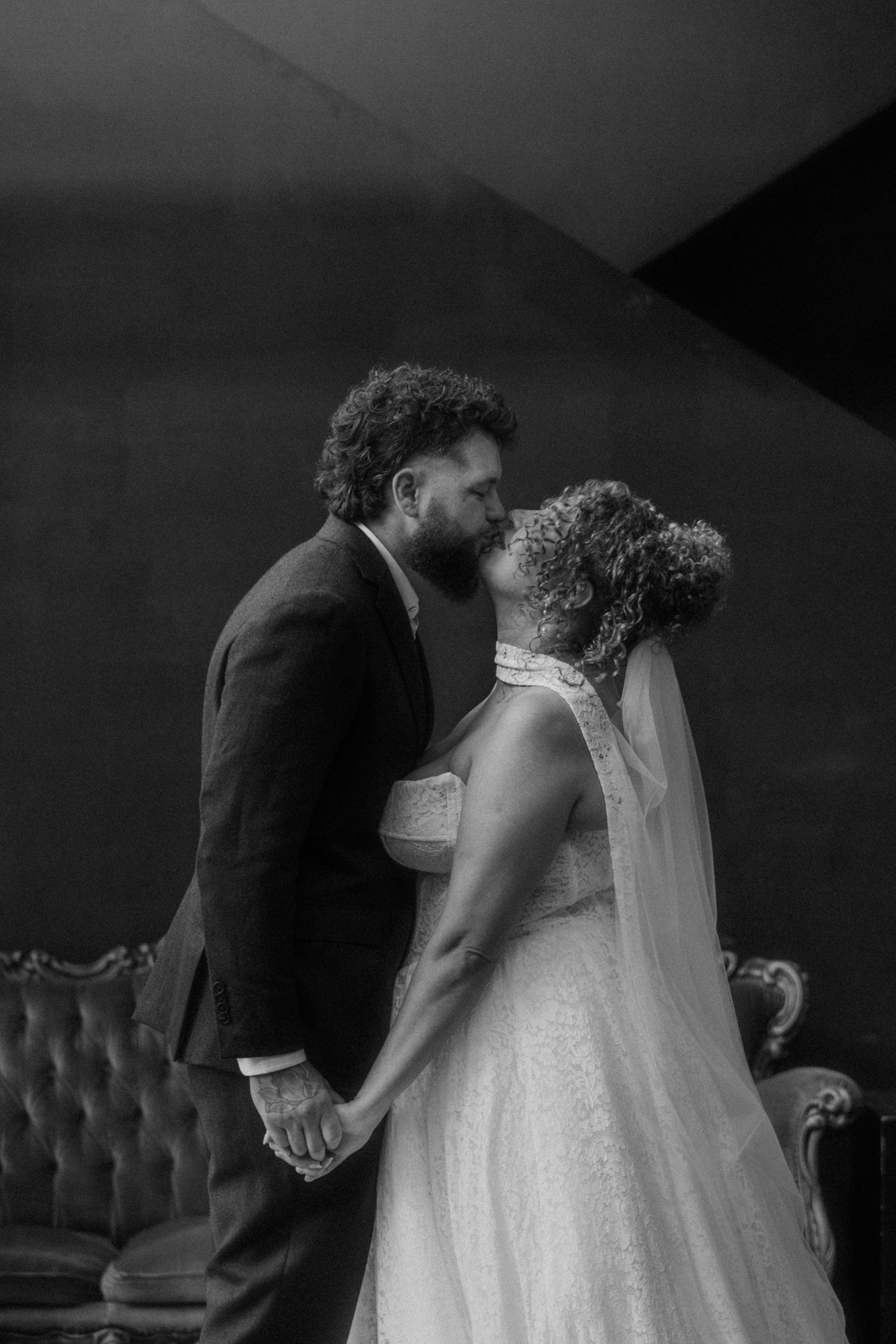 Black and white photo of a bride and groom sharing a kiss while holding hands on their wedding day.