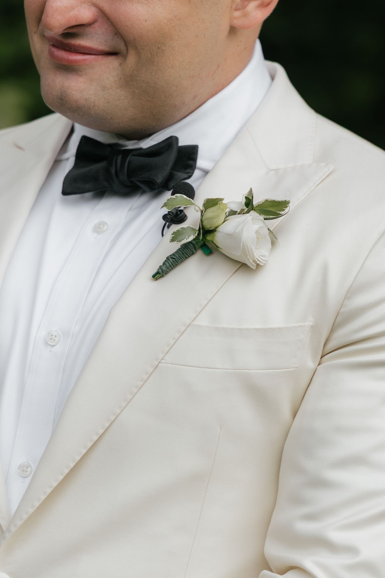 Groom in white tuxedo jacket with black bow tie and white rose boutonniere with green leaves at wedding.