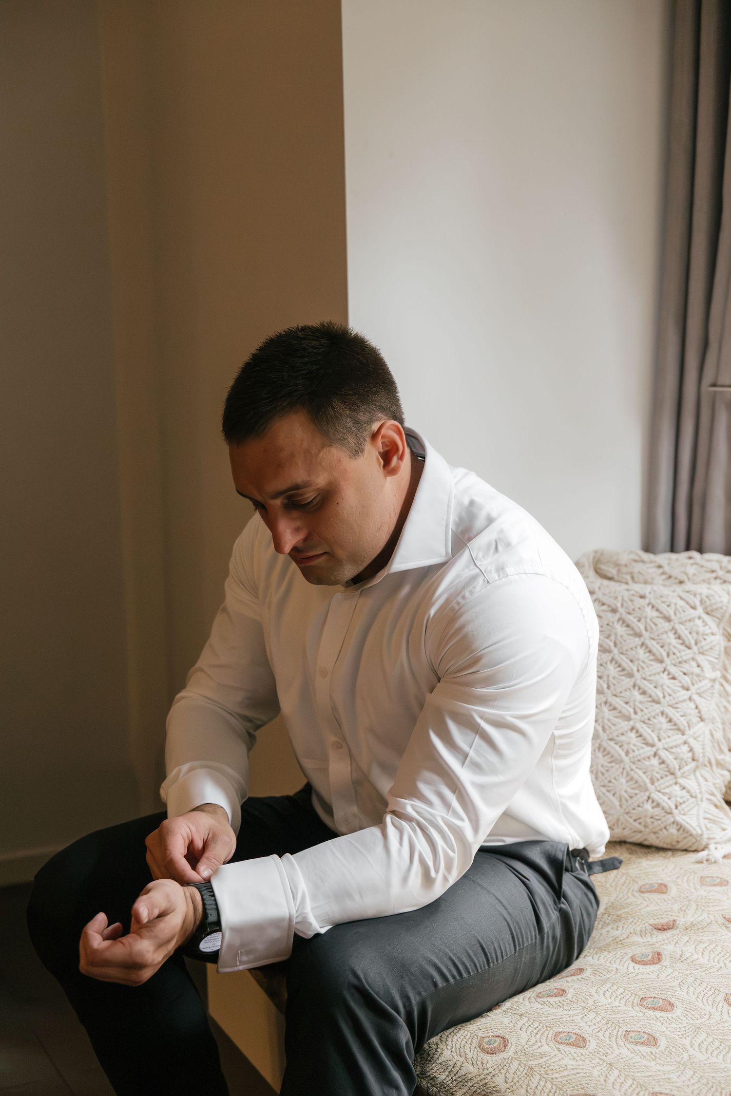 Man in white dress shirt adjusting his watch cufflink while sitting on bed, getting dressed for formal occasion.